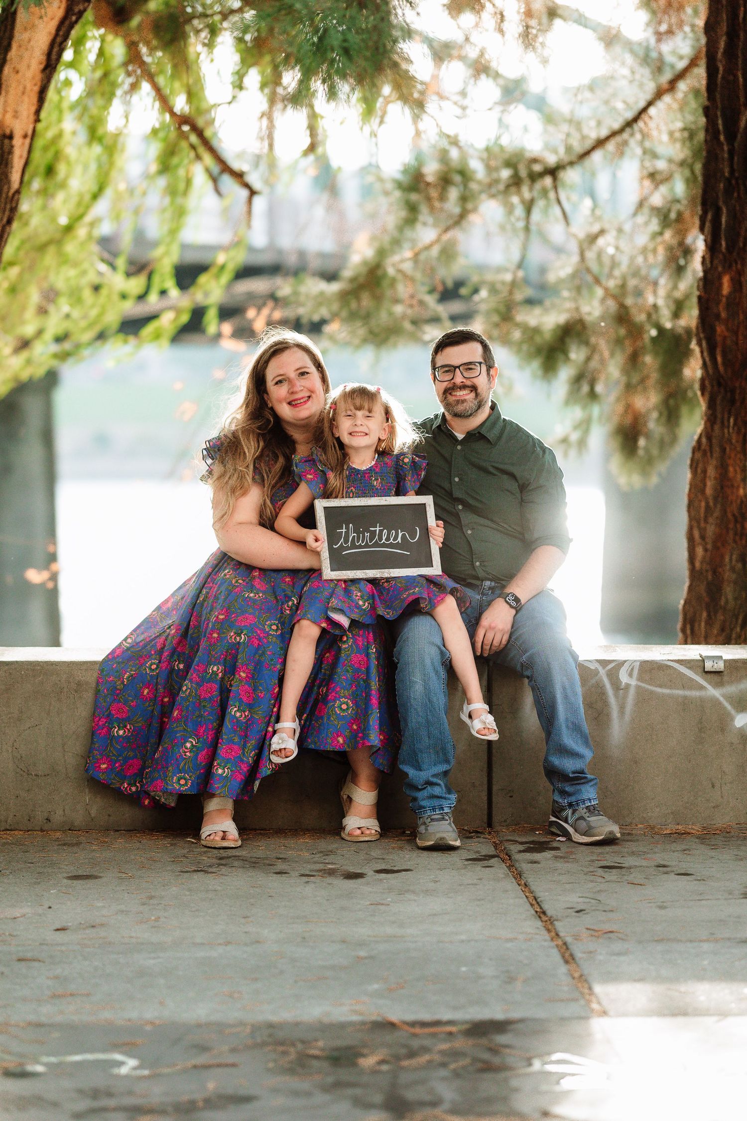 A happy family of three sits outdoors holding a chalkboard sign, with a young girl in a purple floral dress between her parents.