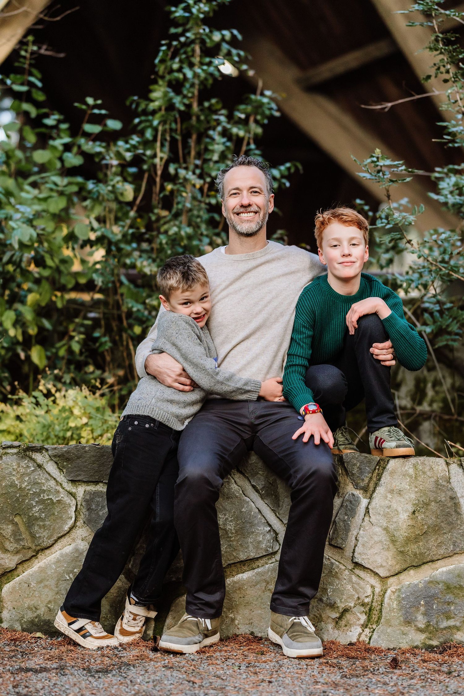 A smiling man sits on a stone wall outdoors with two red-haired boys beside him in a lush green garden setting.