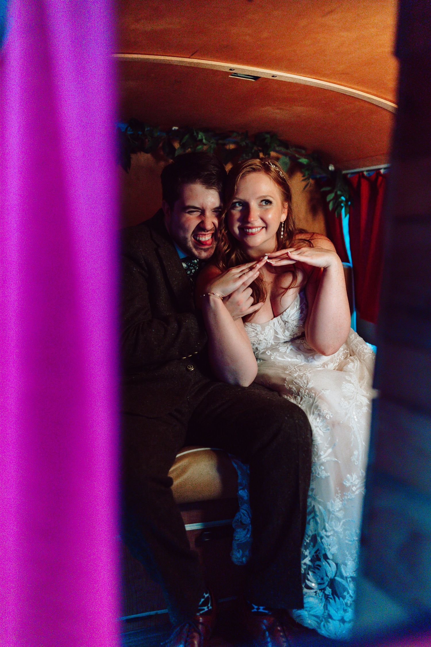 Bride and groom laughing together inside a colorful vintage photo booth on their wedding day.