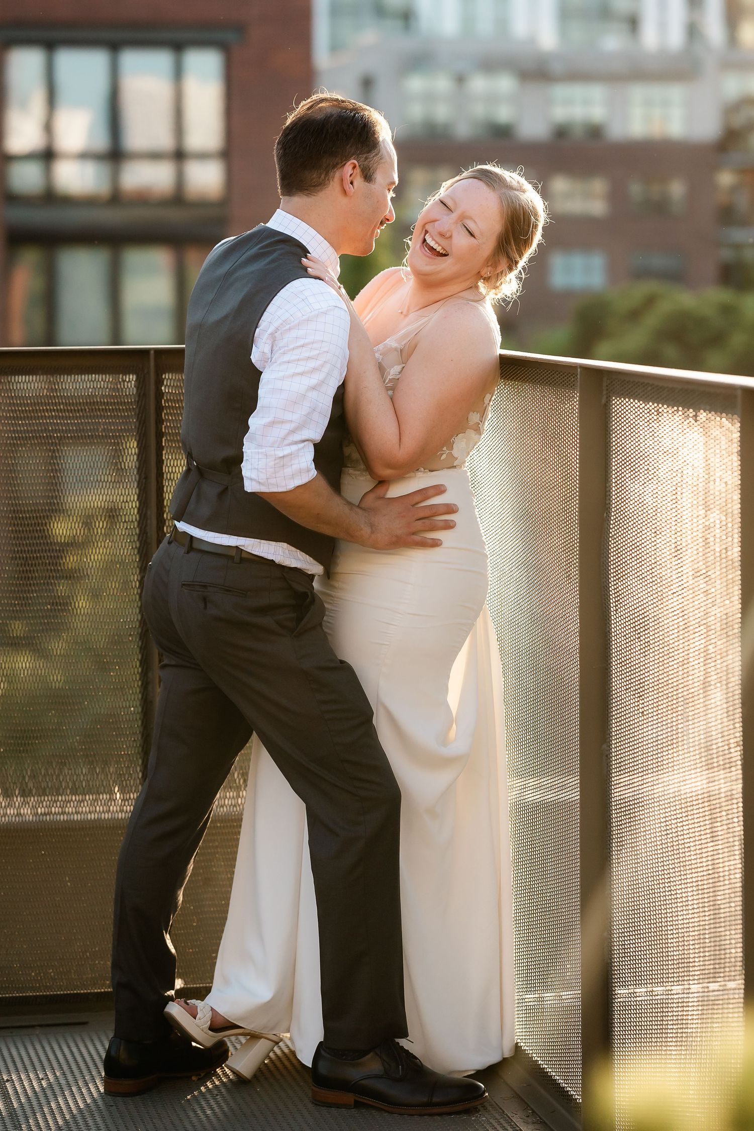 Laughing bride leans against railing as groom embraces her in golden sunset light.