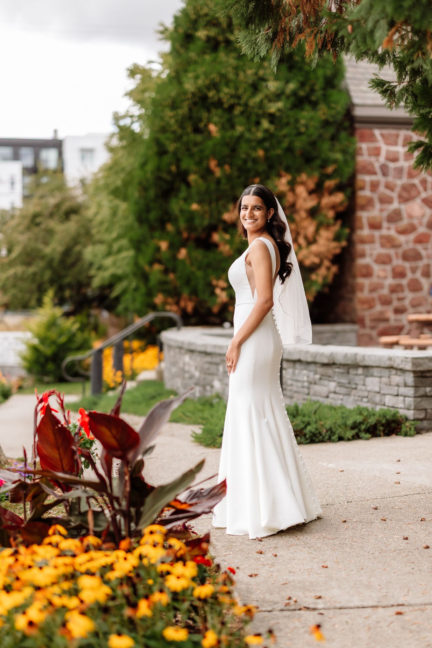 Elegant bride in white fitted gown and veil poses among colorful autumn garden flowers.