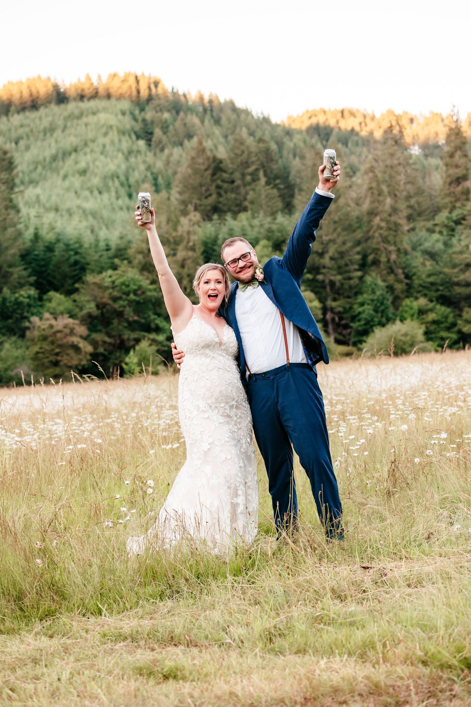 Joyful newlyweds celebrating in a meadow, raising glasses and cheering with forested hills glowing at sunset.