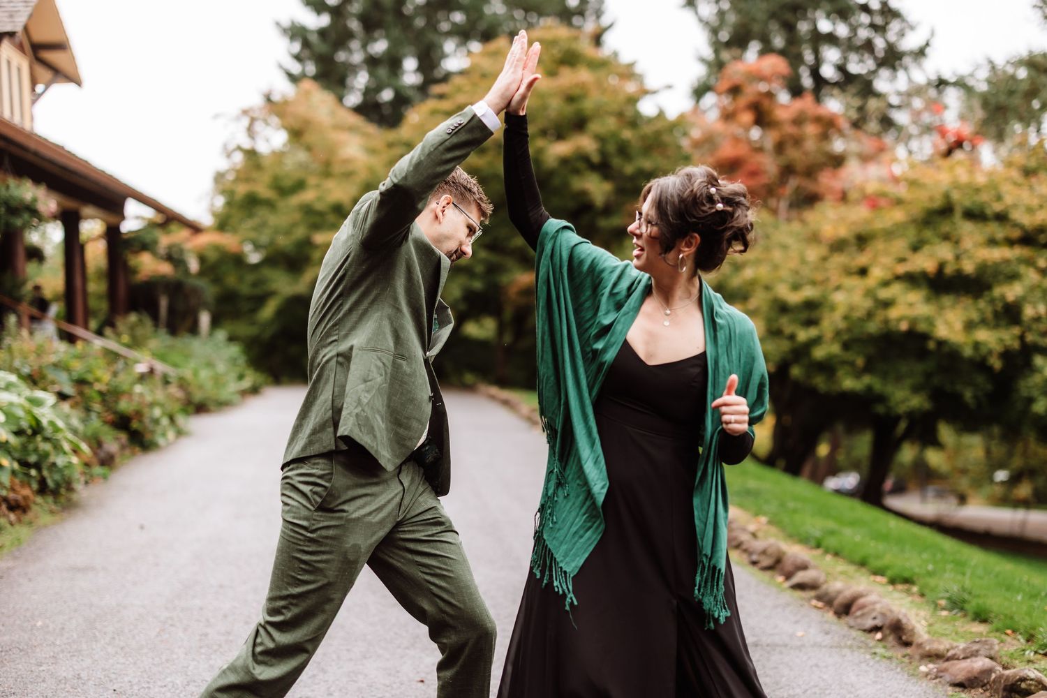 Two wedding guests in green outfits joyfully high-fiving on outdoor garden path.