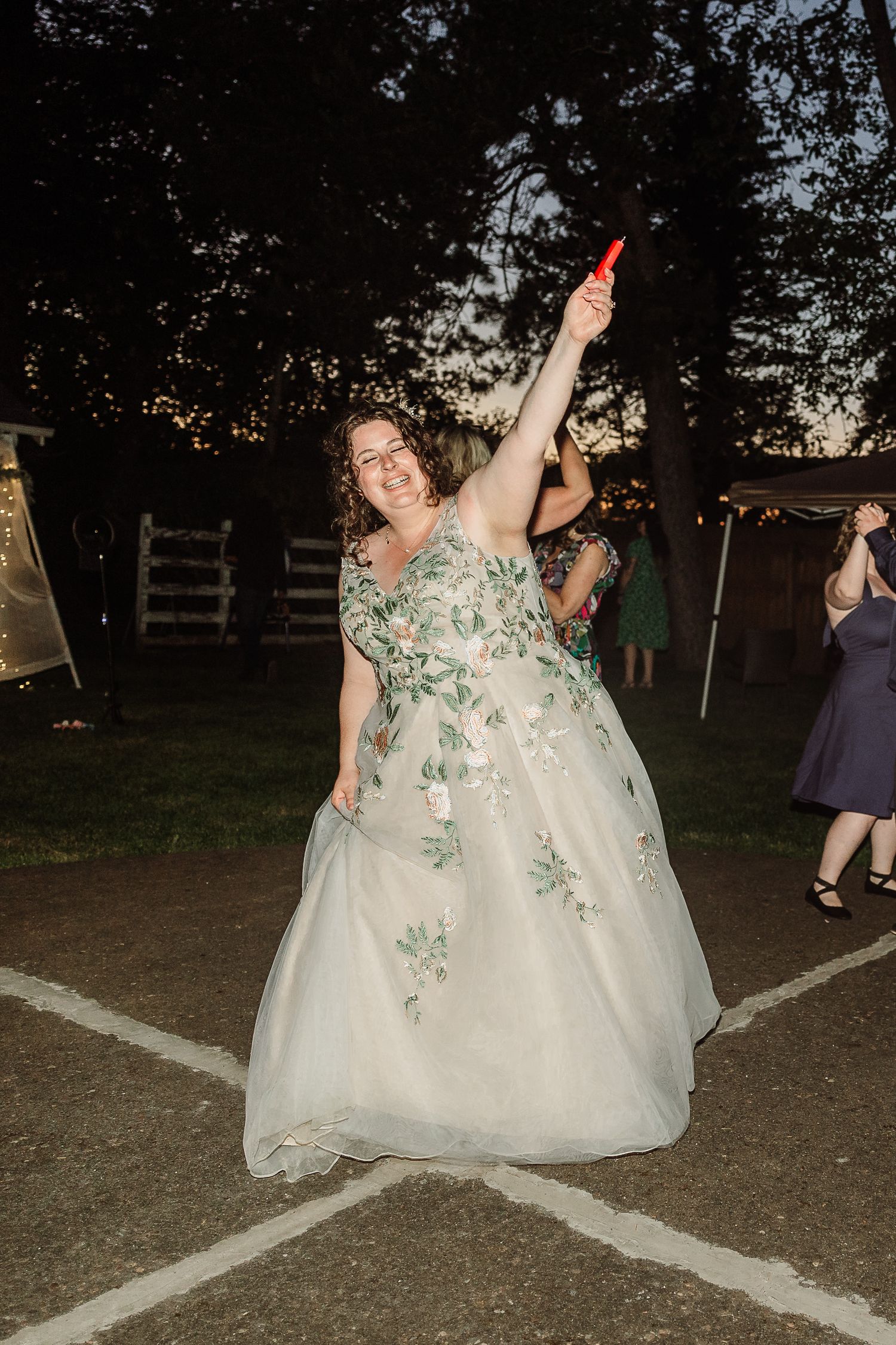 Bride in floral gown raises a red glow stick while dancing outdoors at evening wedding reception.
