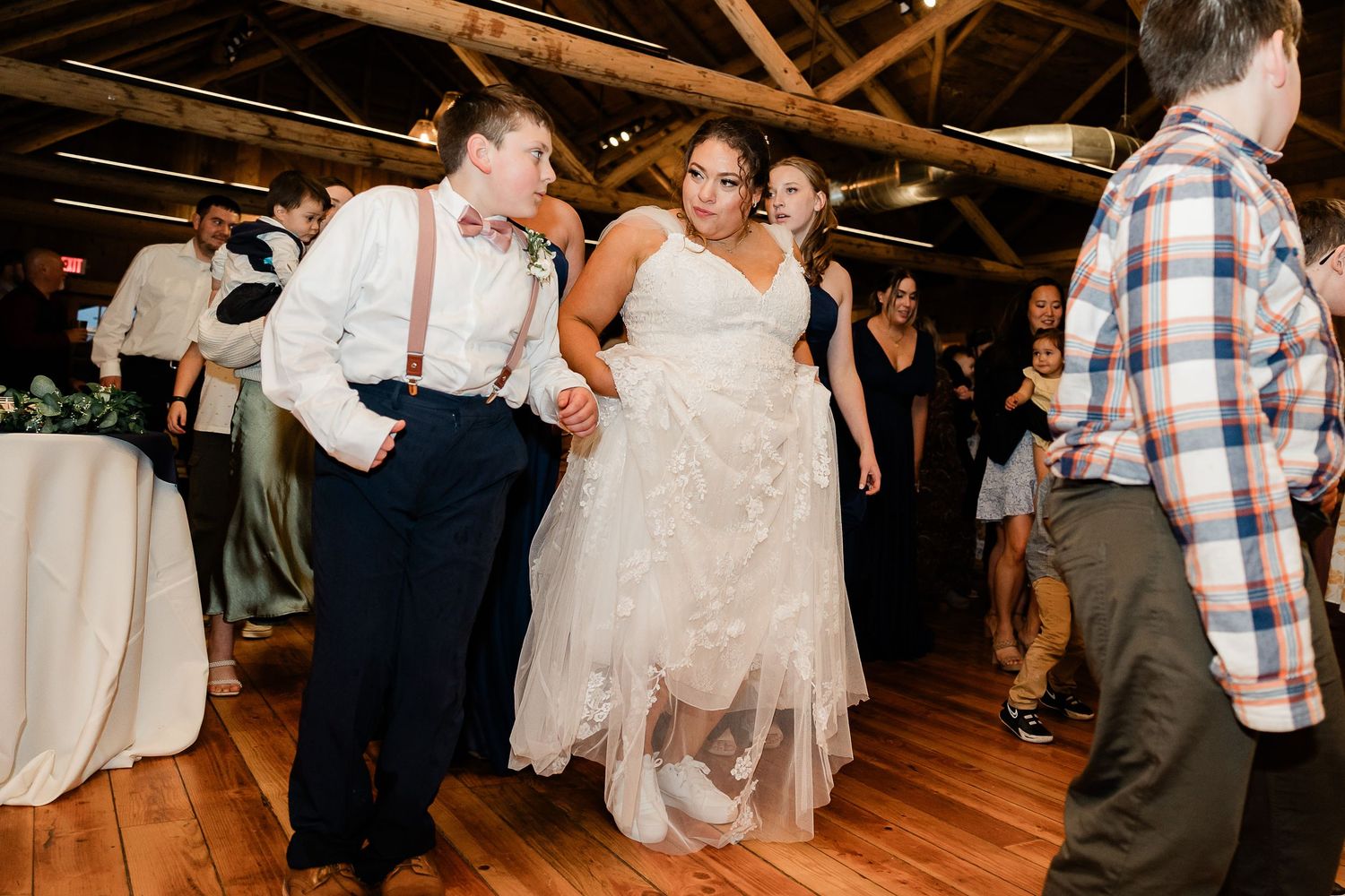 Bride and groom dance energetically with wedding guests on wooden dance floor in rustic barn venue.