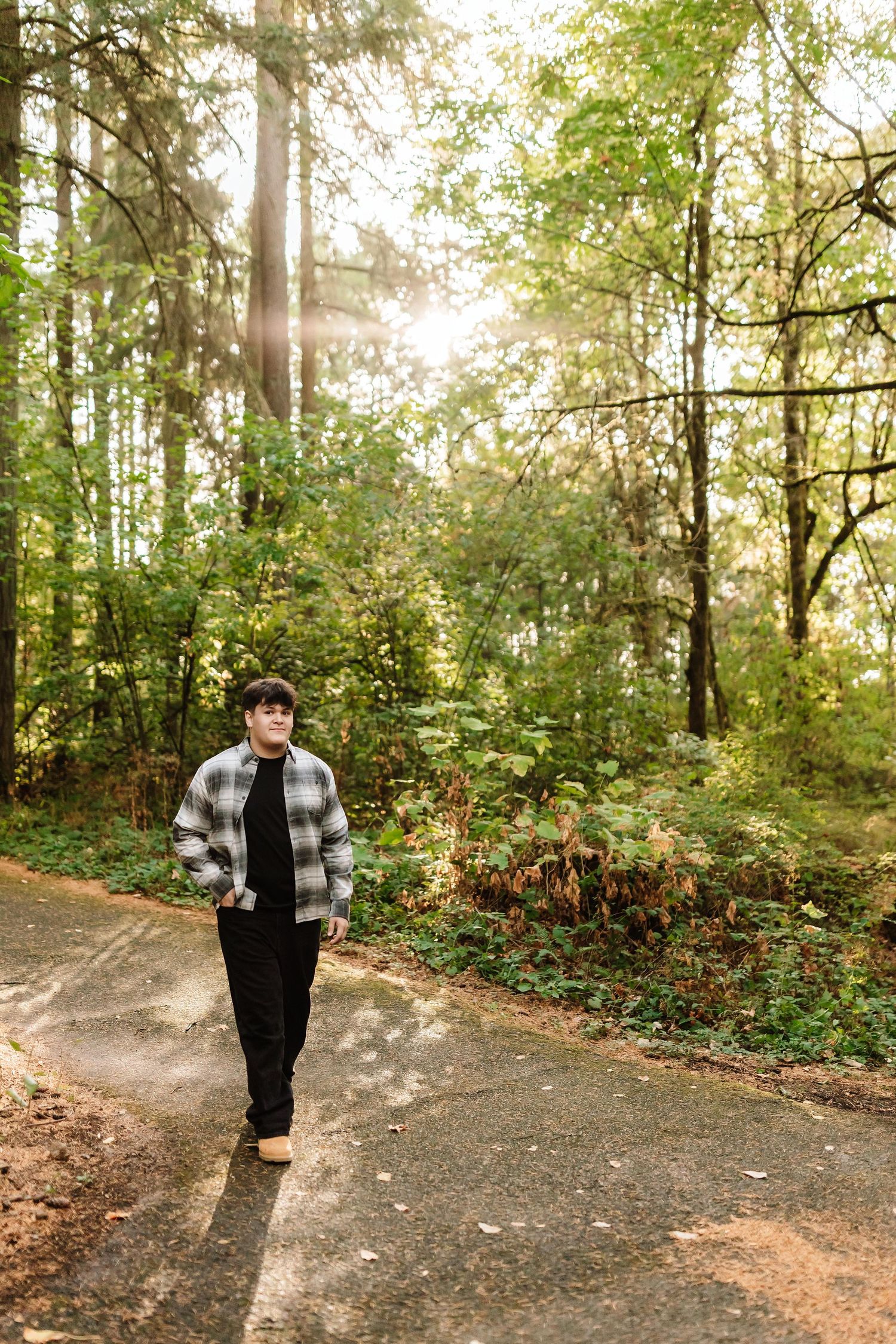 Teen boy in plaid jacket walking on a sunlit forest path among tall green trees.