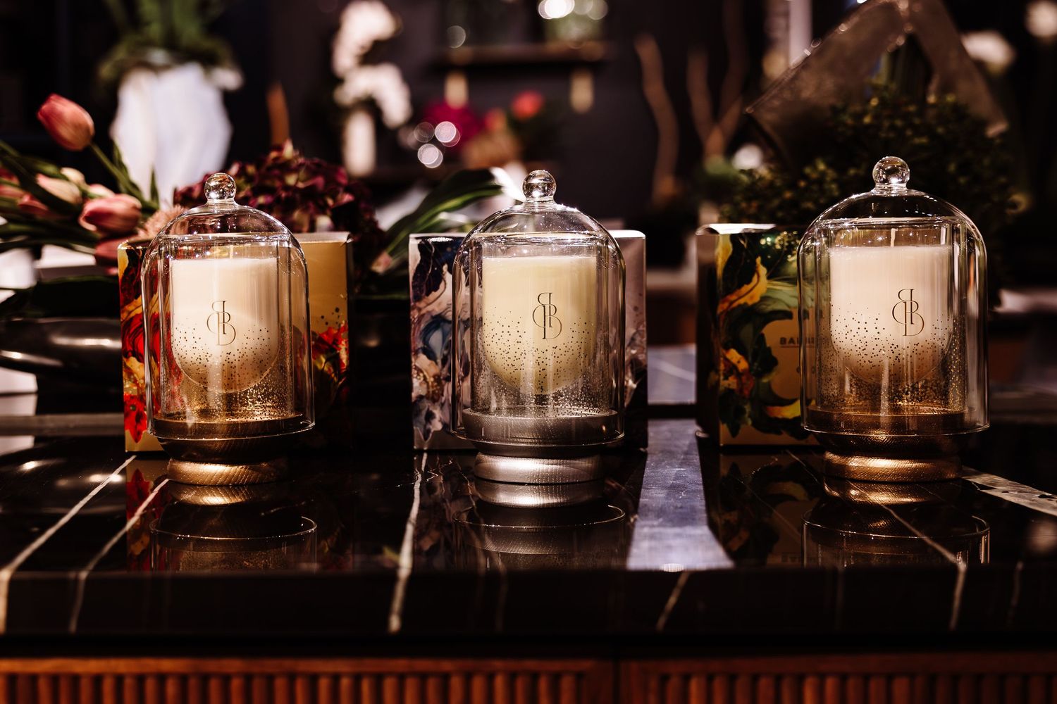 Three elegant glass cloche candles with gold monogram bases displayed on a dark marble surface.
