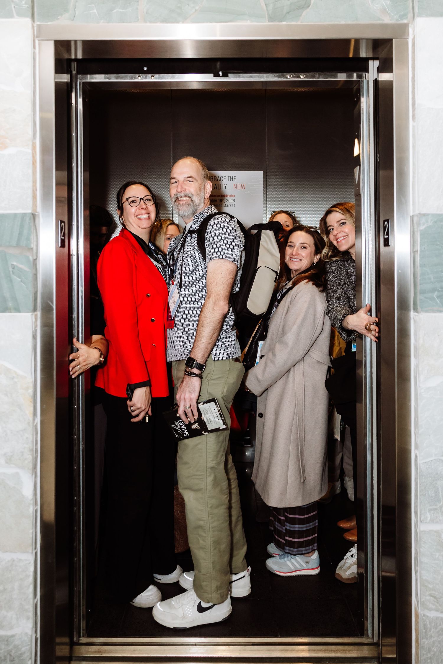 A group of five people smile together inside an elevator at a trade show.