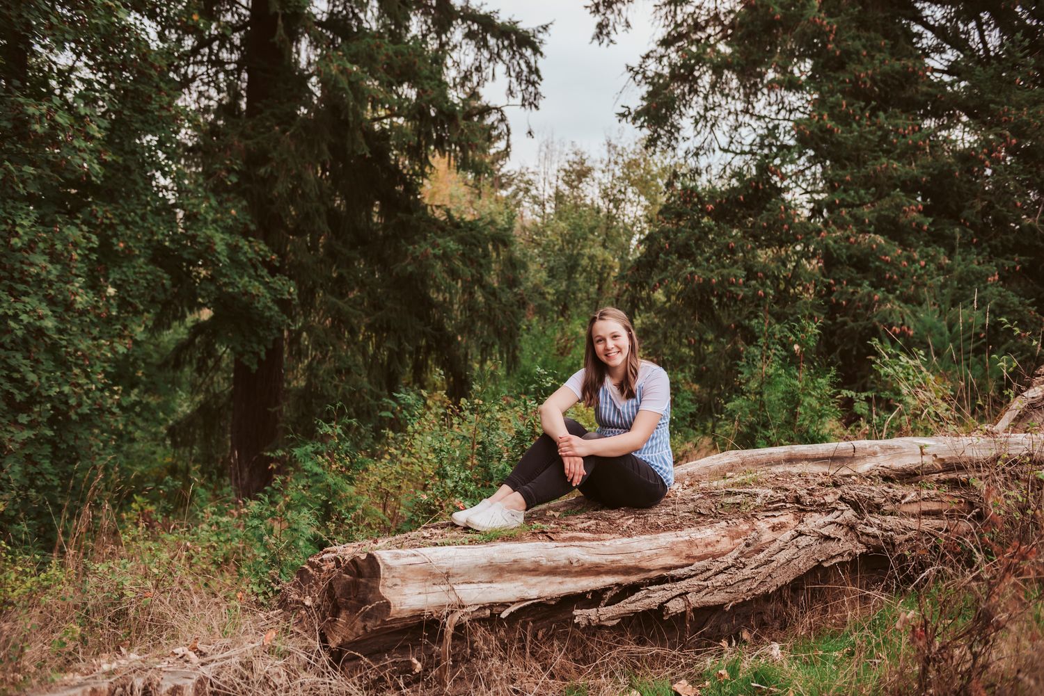 Young woman sitting on a fallen log in a lush green forest, smiling casually in jeans and a blue top.