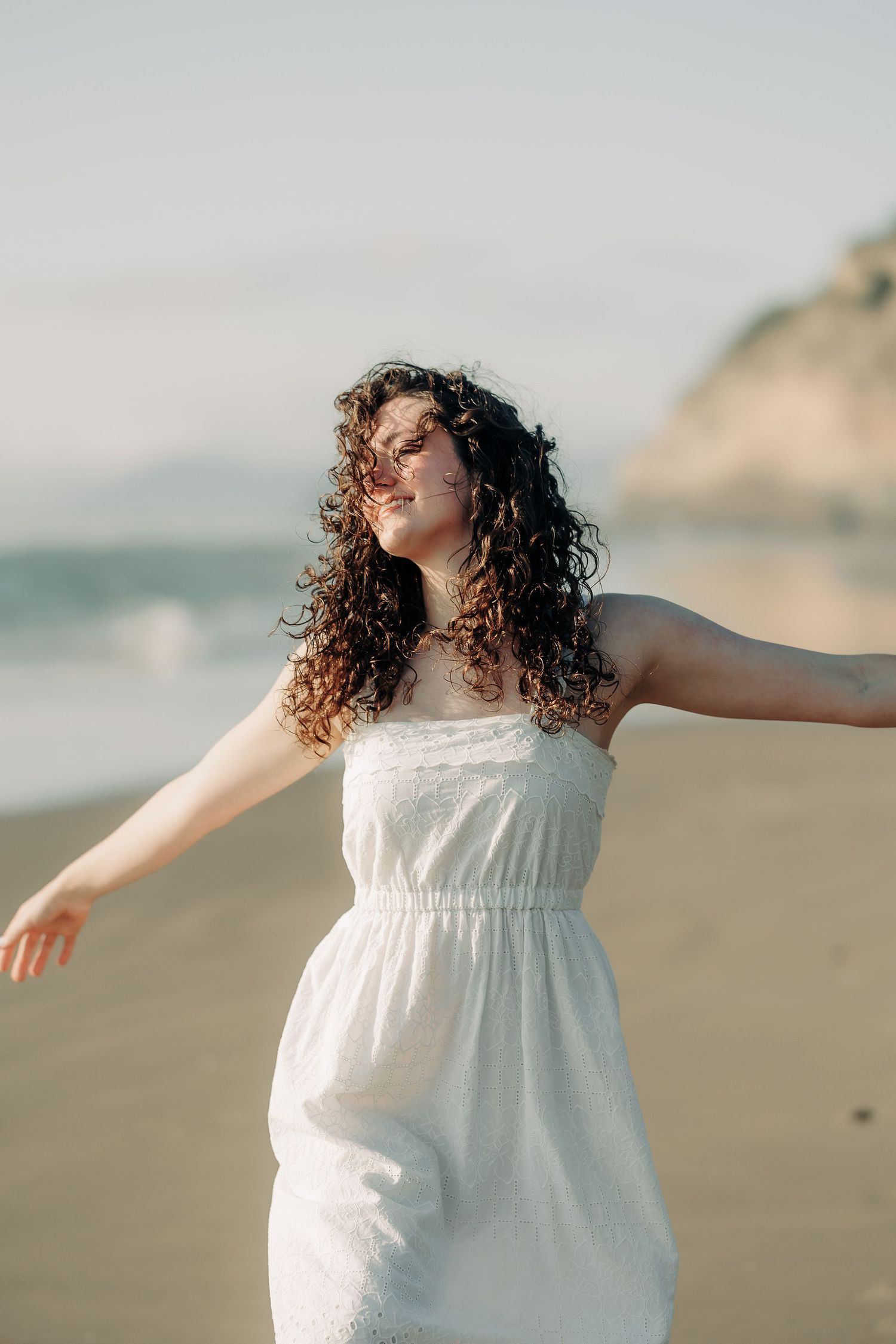 Young woman in white sundress with arms outstretched on a beach at golden hour.