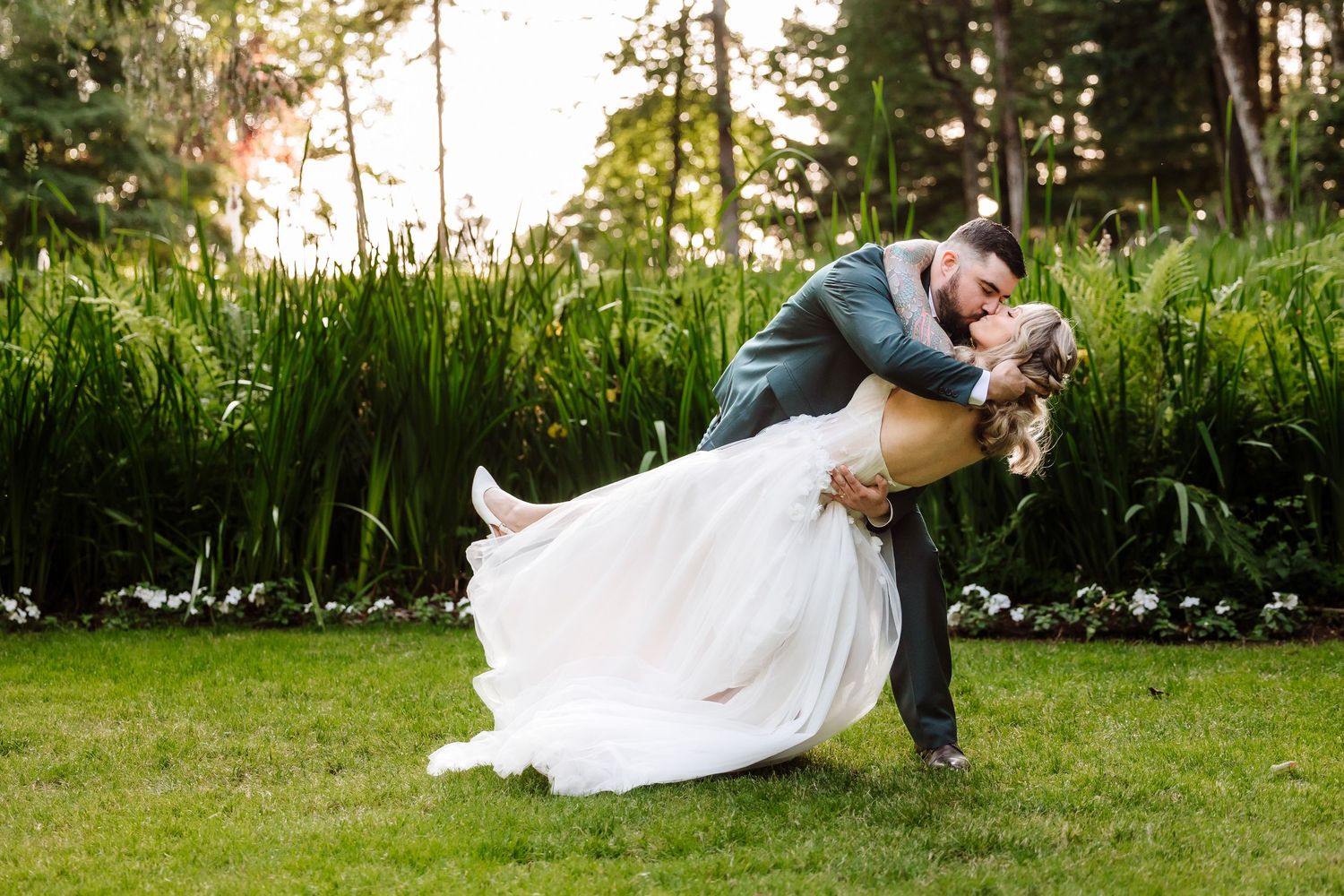 Groom dips bride in flowing white wedding dress on green lawn surrounded by tall garden plants.