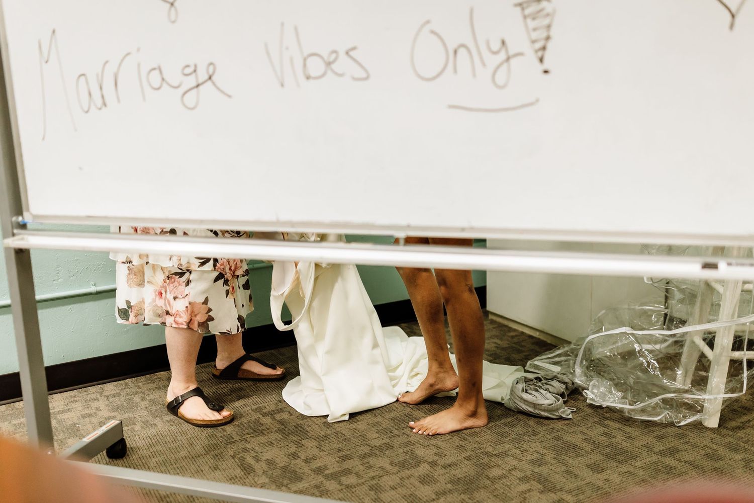 Bride in white wedding gown standing barefoot near a whiteboard with wedding messages.