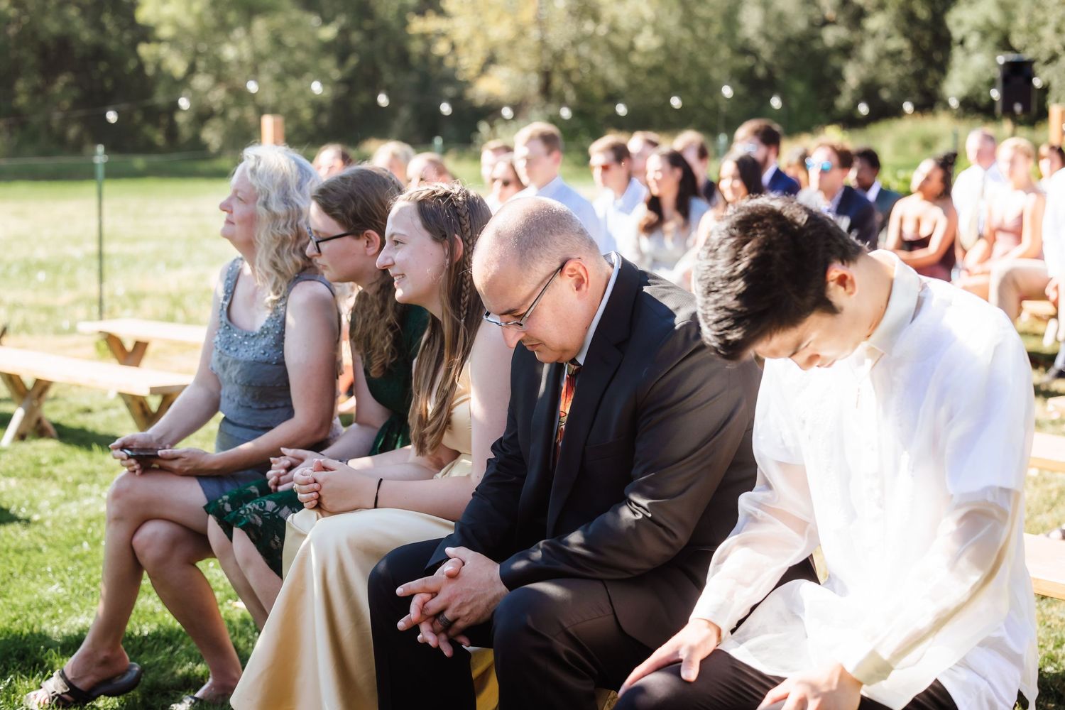 Wedding guests seated on wooden benches during outdoor garden ceremony on sunny afternoon.
