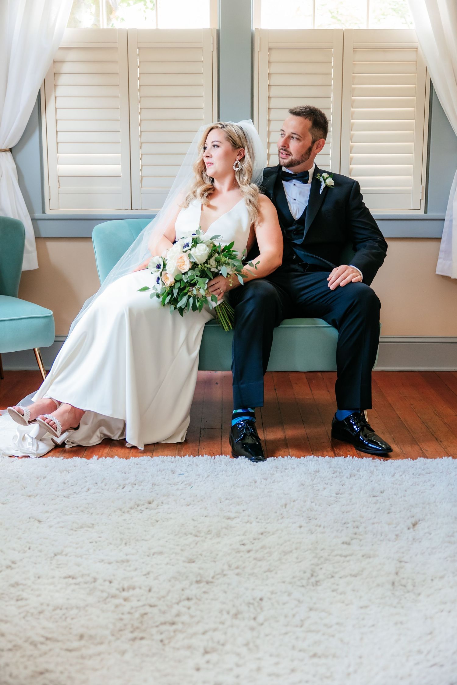 Bride and groom sitting together on a teal bench indoors, bride holding a white bouquet in wedding attire.