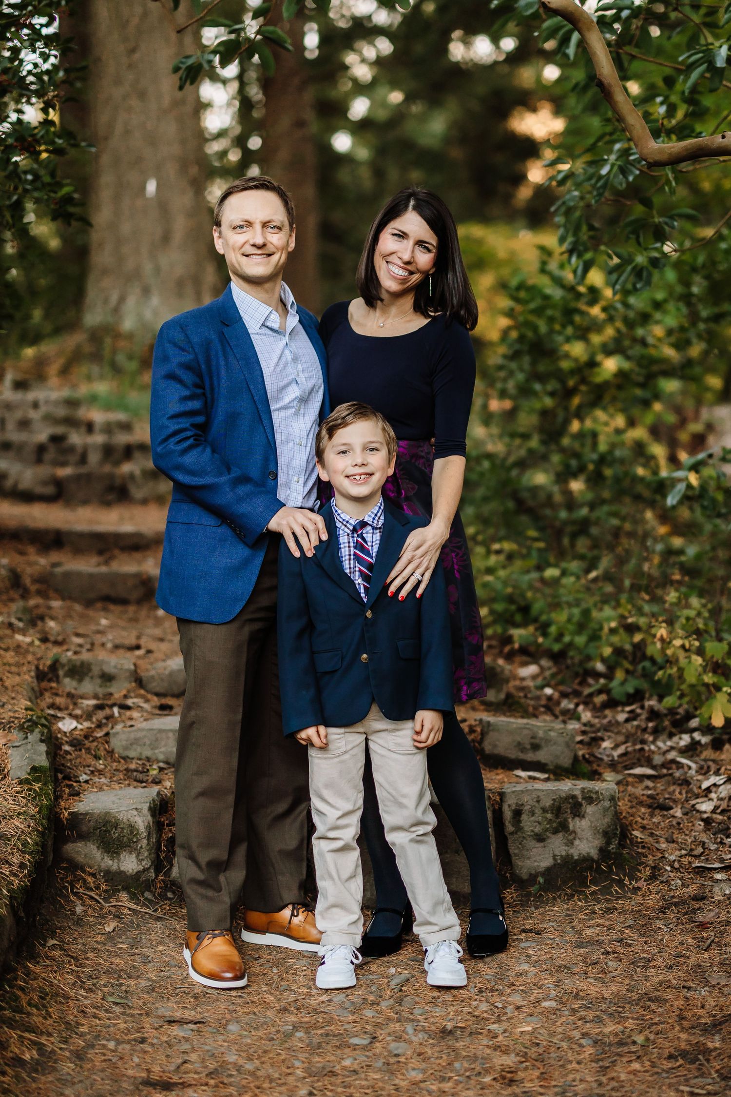 Family of three posing outdoors in a wooded garden, parents and young boy dressed in coordinating navy blue outfits.