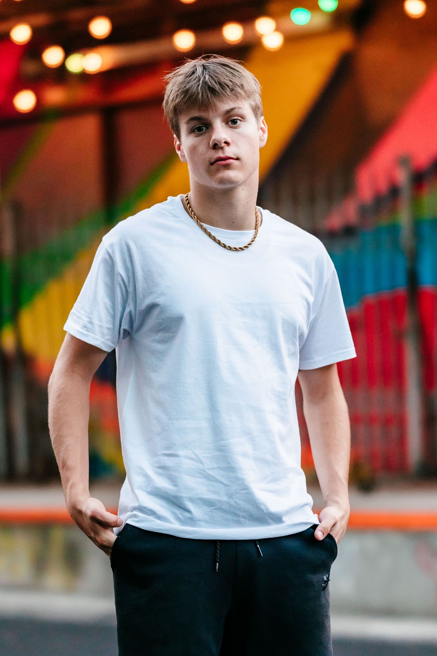 Young man in white t-shirt and gold chain poses confidently outdoors with colorful graffiti wall and string lights behind him.