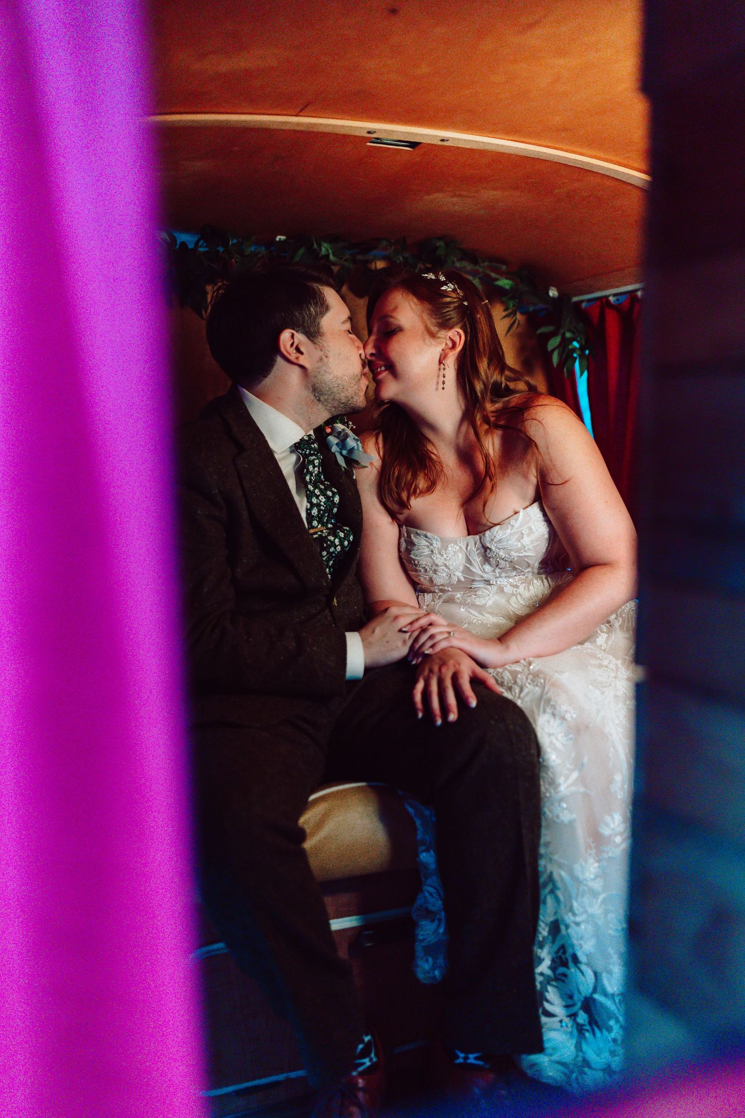 Bride and groom share a romantic kiss inside a cozy vintage photo booth with colorful curtains.