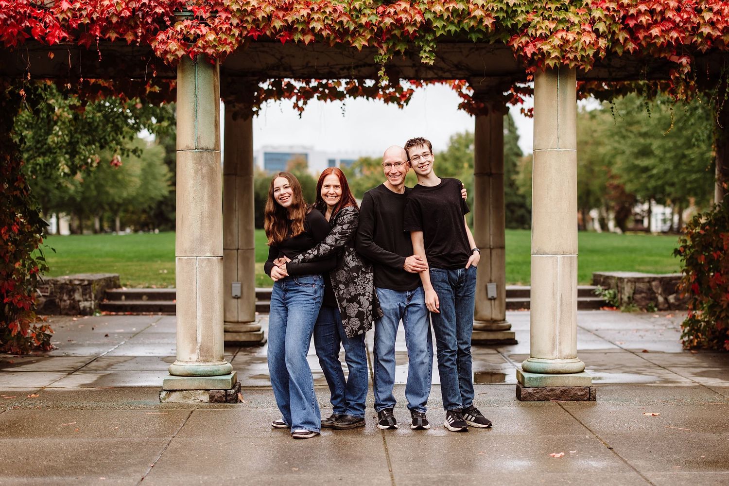 Family of four smiling under a red autumn vine pergola in an outdoor fall portrait.