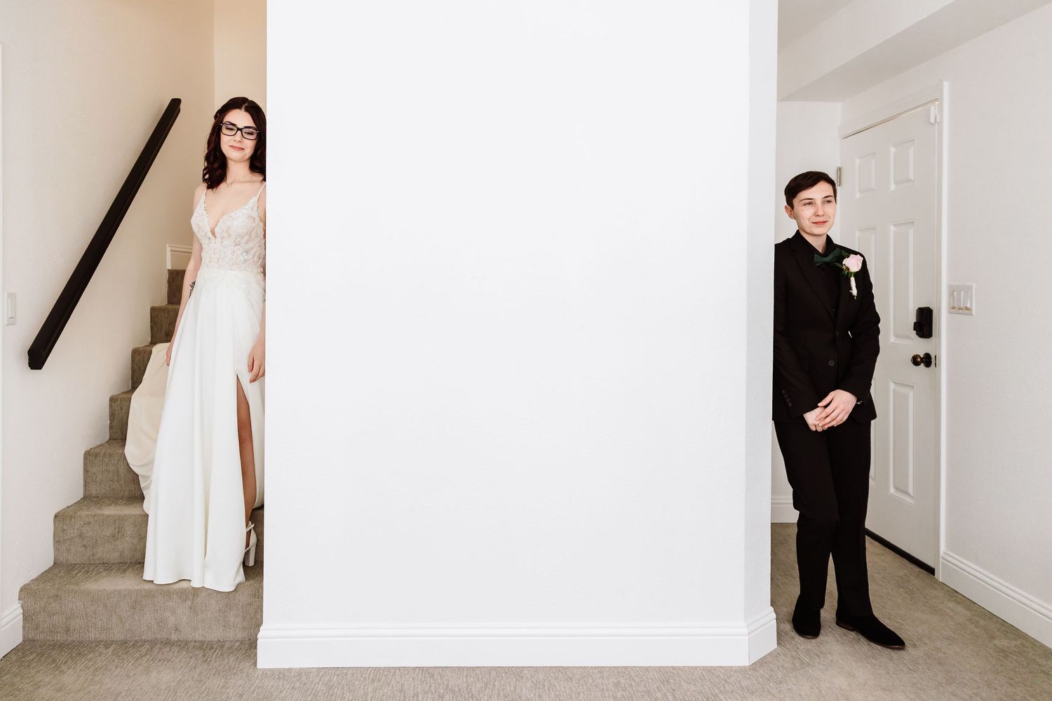 Bride in white gown and groom in black suit stand on opposite sides of white wall.