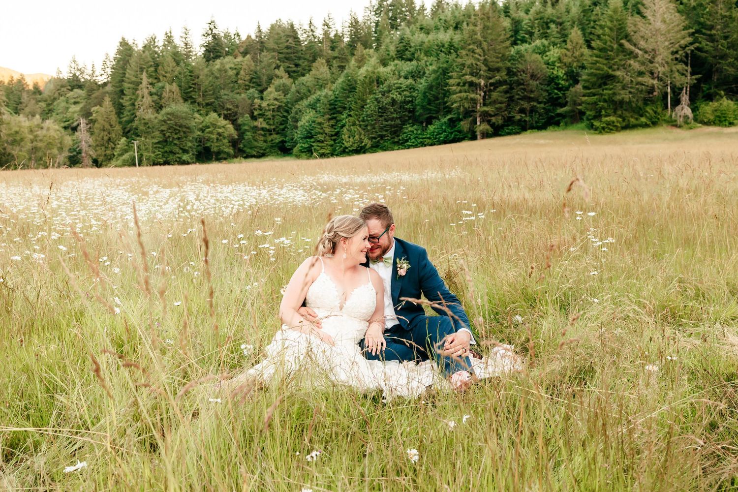 Bride and groom sitting romantically in a wildflower meadow with lush green forest backdrop on their wedding day.