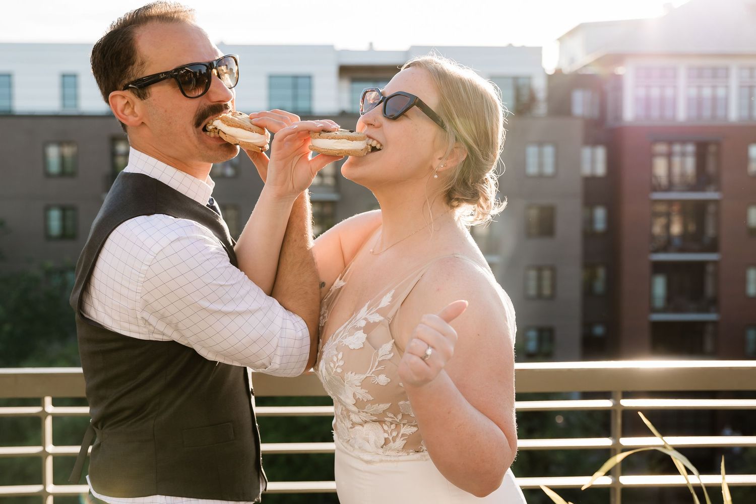 Playful bride and groom share a bite of food on sunny rooftop terrace.