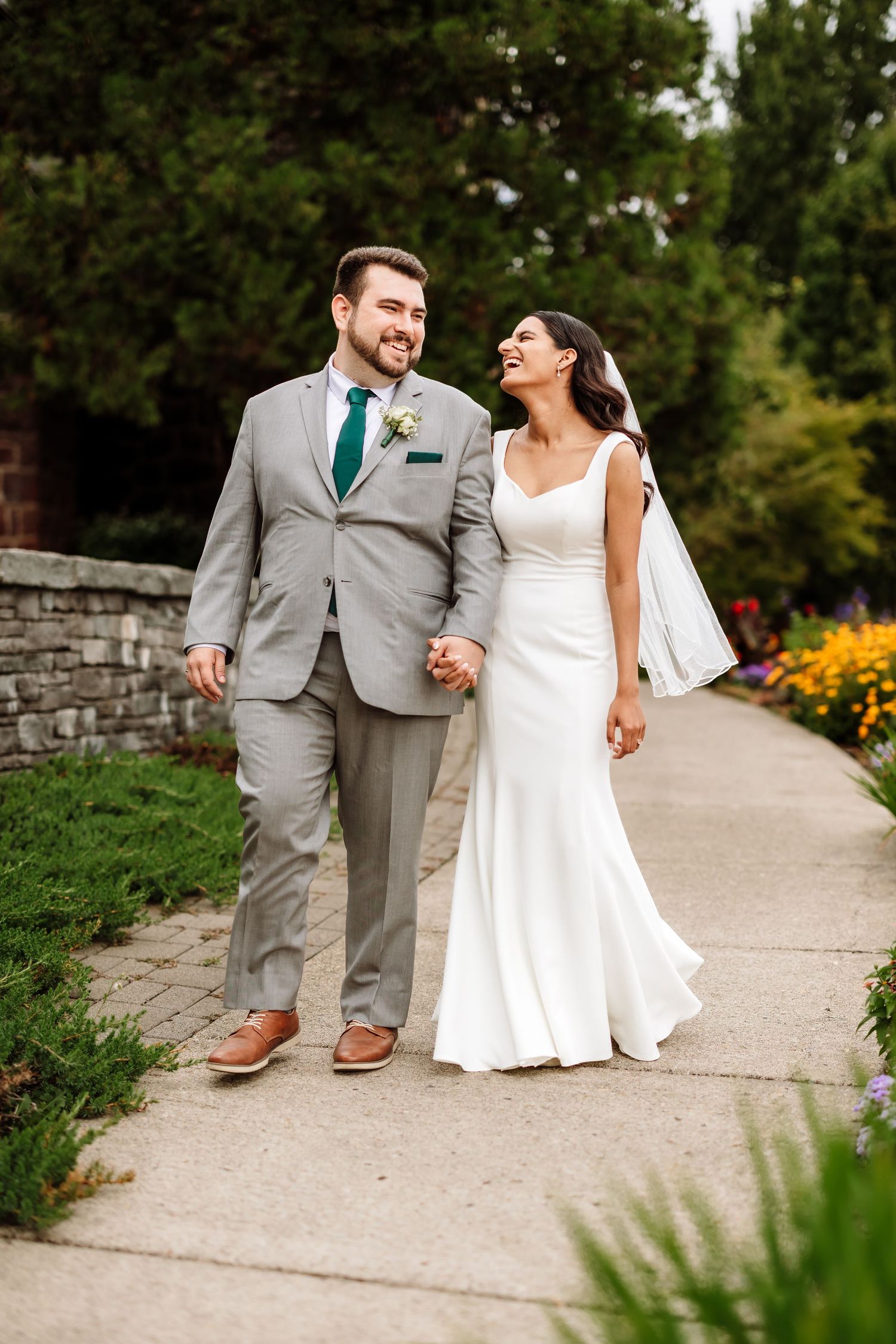 Smiling bride and groom walk hand-in-hand along garden path in wedding attire.