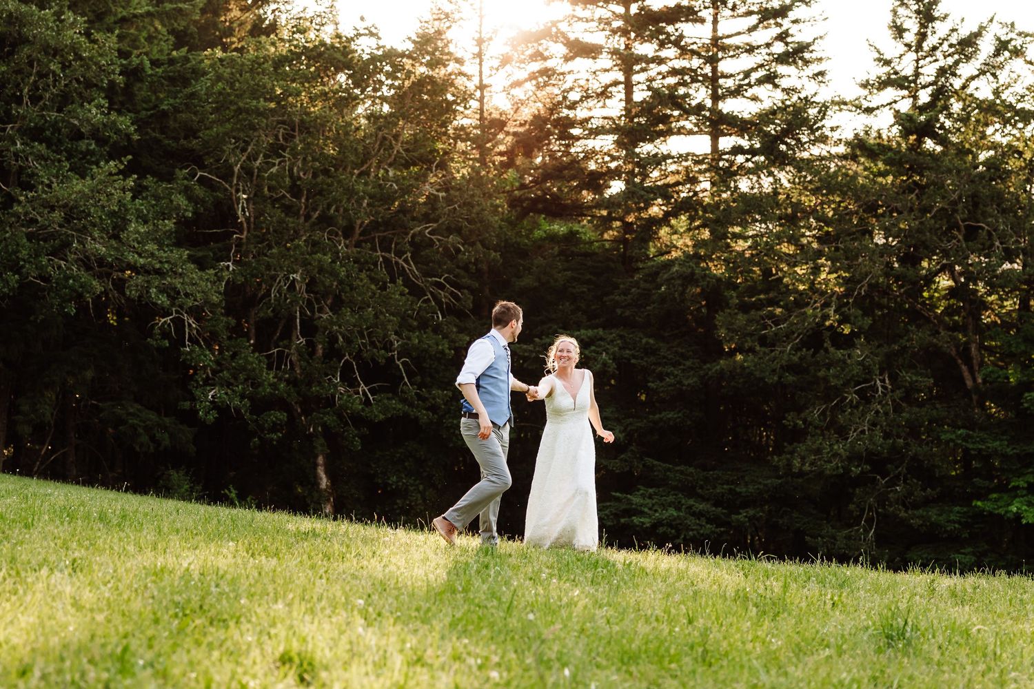Bride and groom walk hand in hand across a sunlit green meadow with pine trees behind.
