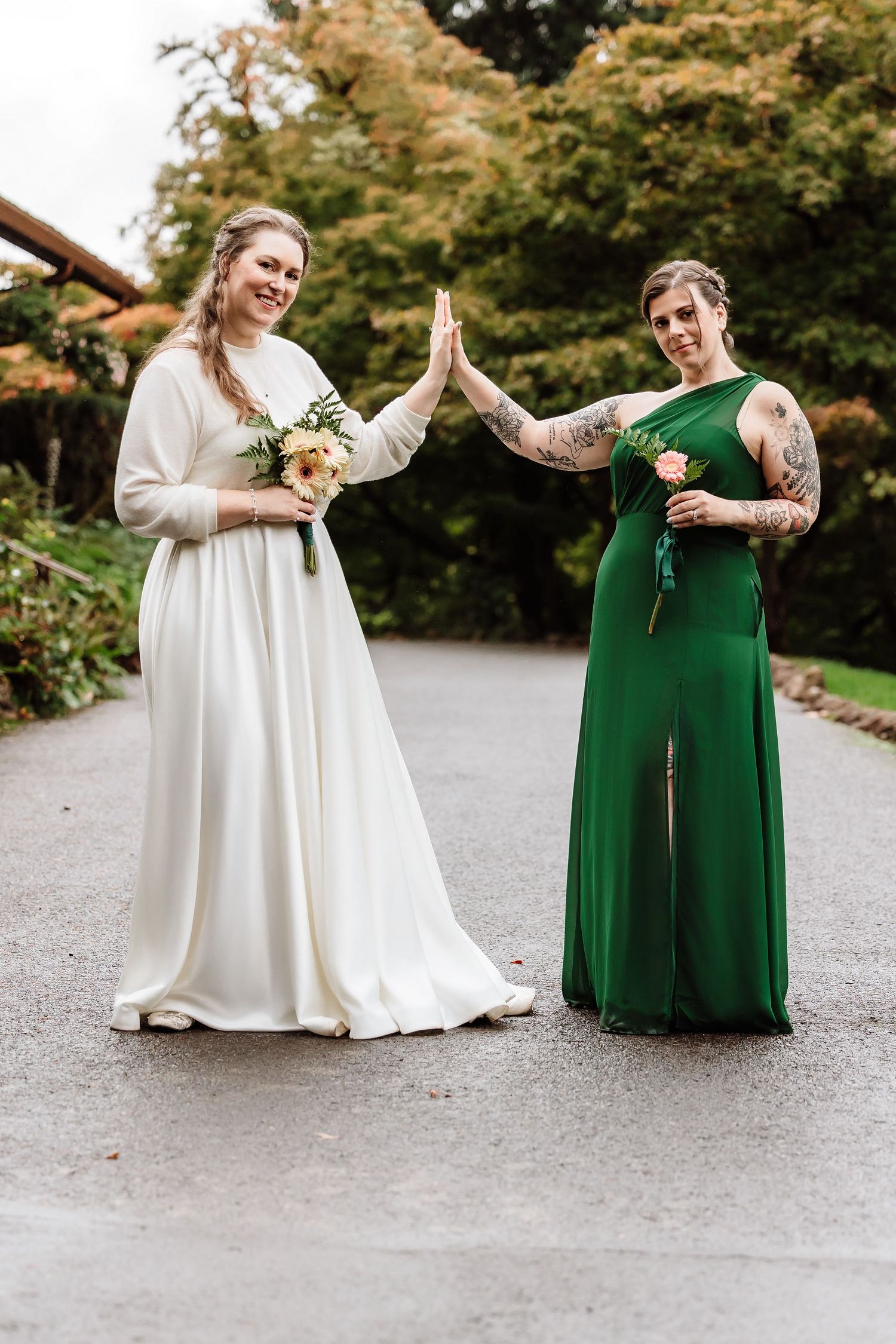 Bride in white gown and bridesmaid in green dress giving high five outdoors at wedding.