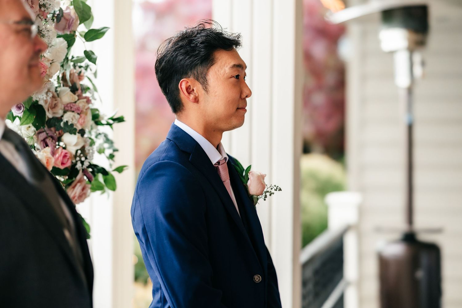 Groom in navy suit waits anxiously at altar adorned with pink floral arrangements during ceremony.
