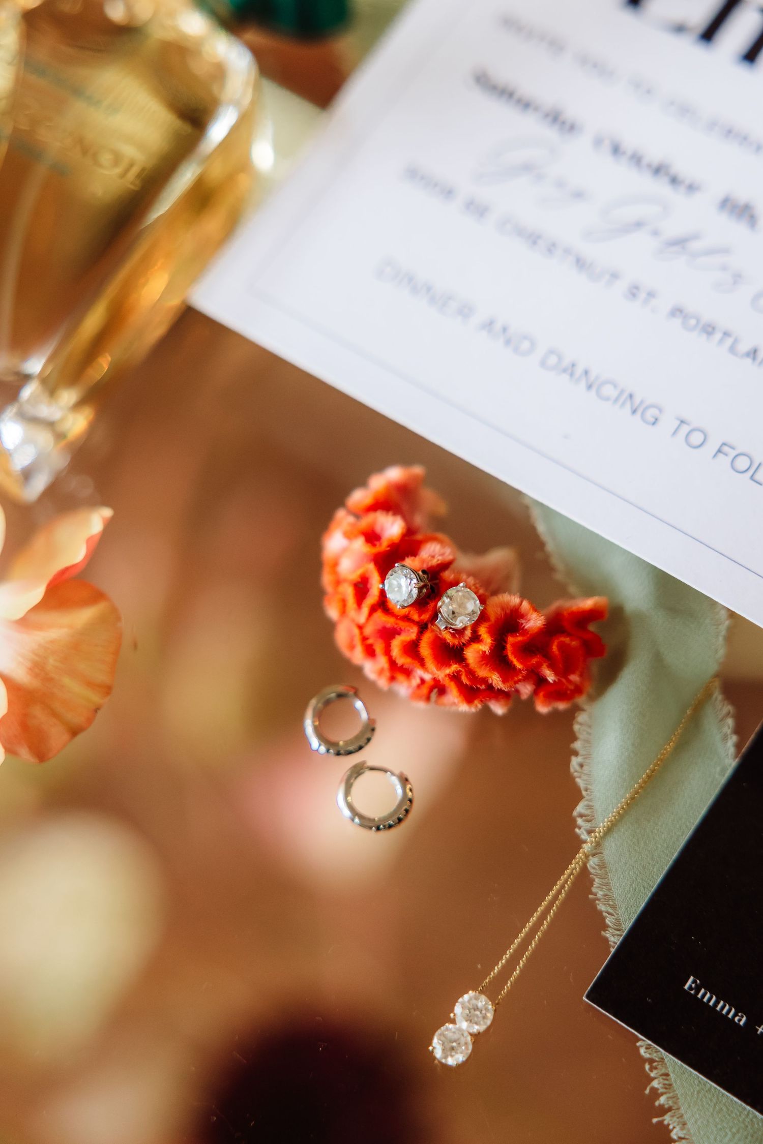Elegant wedding jewelry flat lay with diamond earrings, rings, and necklace on red lace beside an invitation.