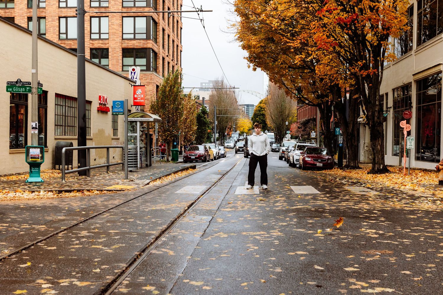 Person crossing a street lined with autumn trees and tram tracks in a vibrant urban neighborhood on a cloudy fall day.