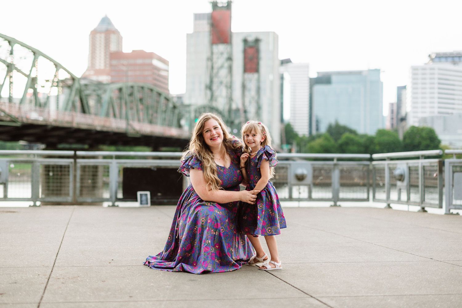 Mother and daughter in matching purple dresses pose together on a waterfront plaza with a city bridge and skyline behind them.