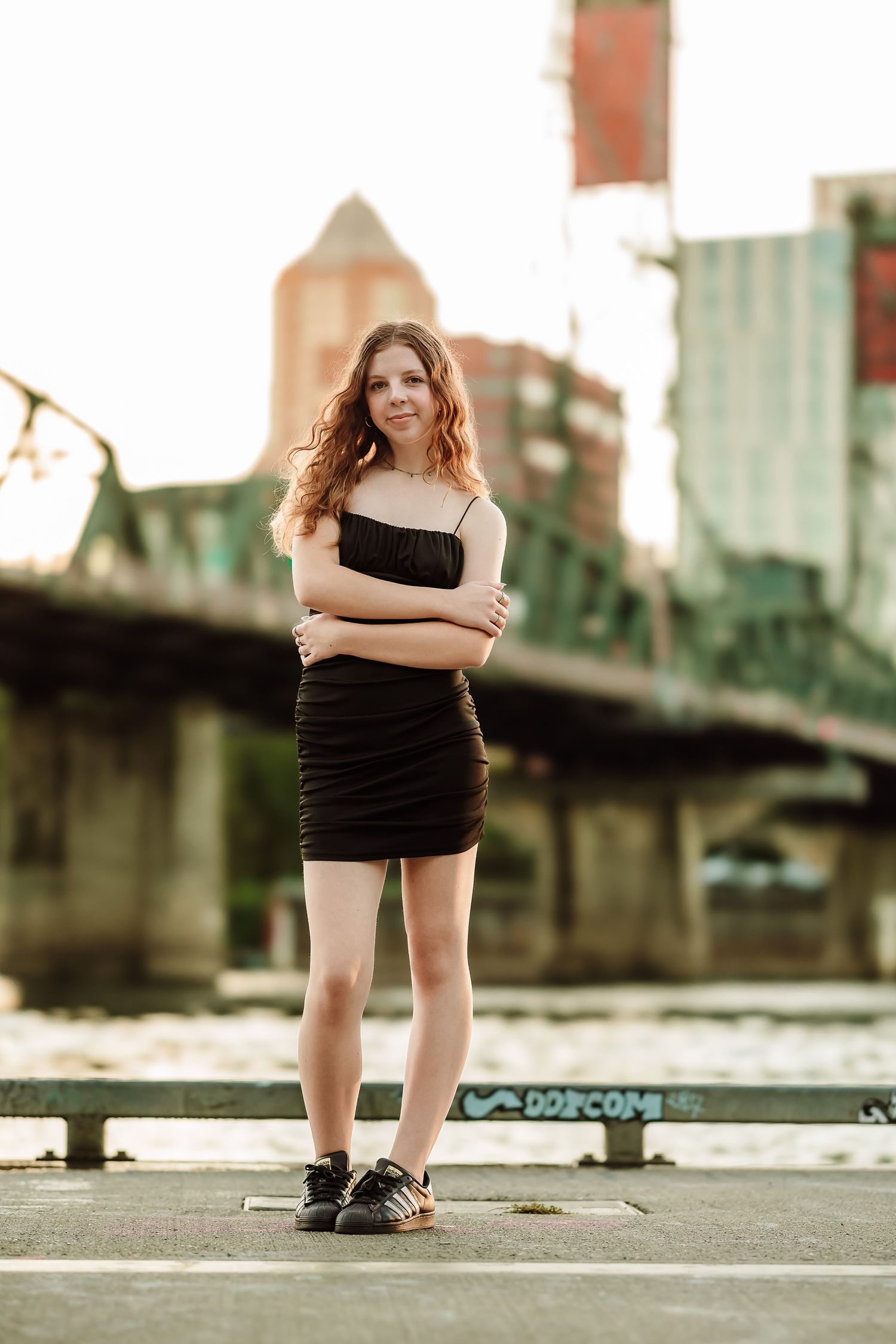 Young woman in black mini dress with crossed arms poses confidently on urban waterfront with bridge and city buildings behind her.