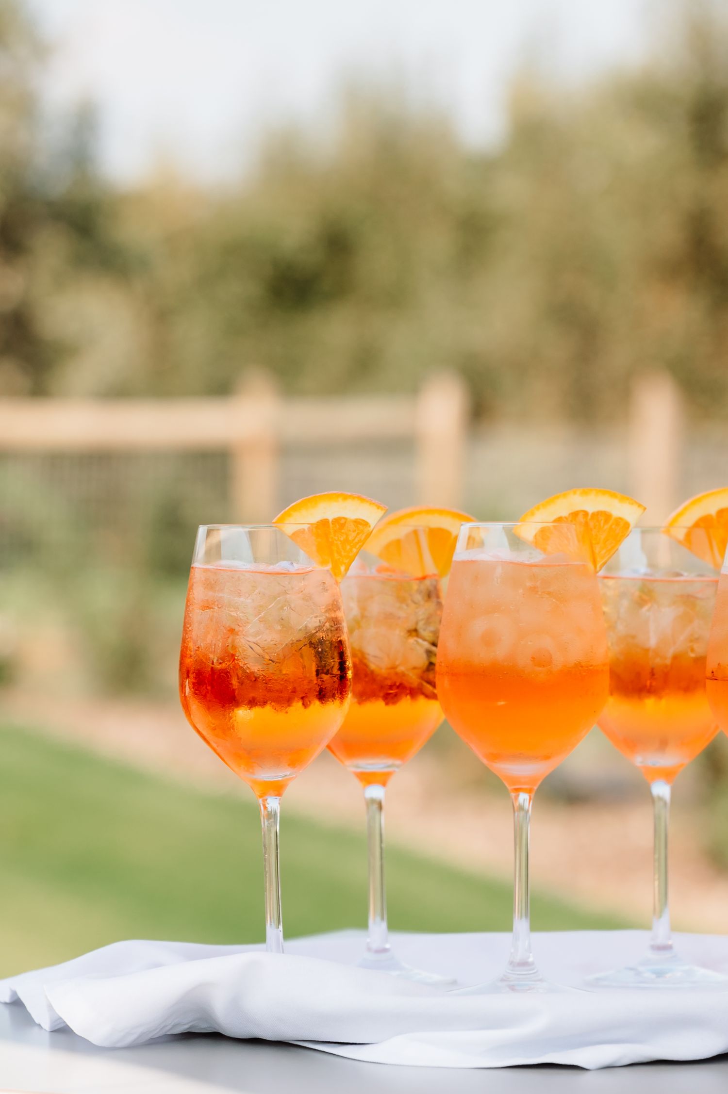 Four Aperol Spritz cocktails garnished with orange slices lined up on a white cloth outdoors.
