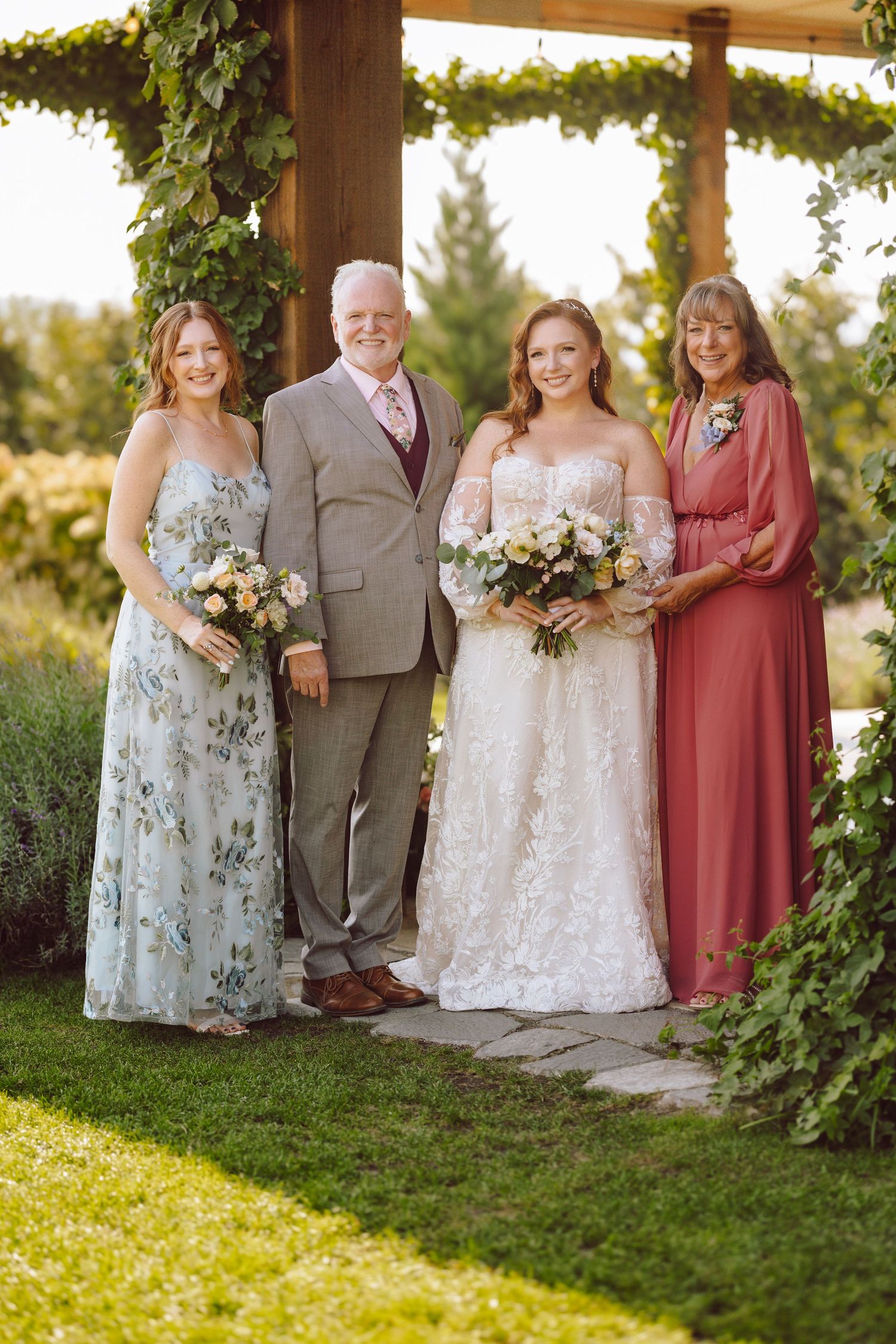 Bride in lace gown poses with family under a vine-covered pergola in a garden wedding setting.