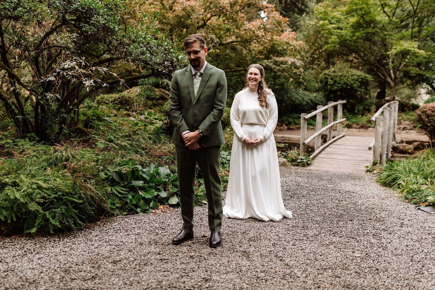 Bride and groom stand together on gravel path surrounded by lush green garden foliage.