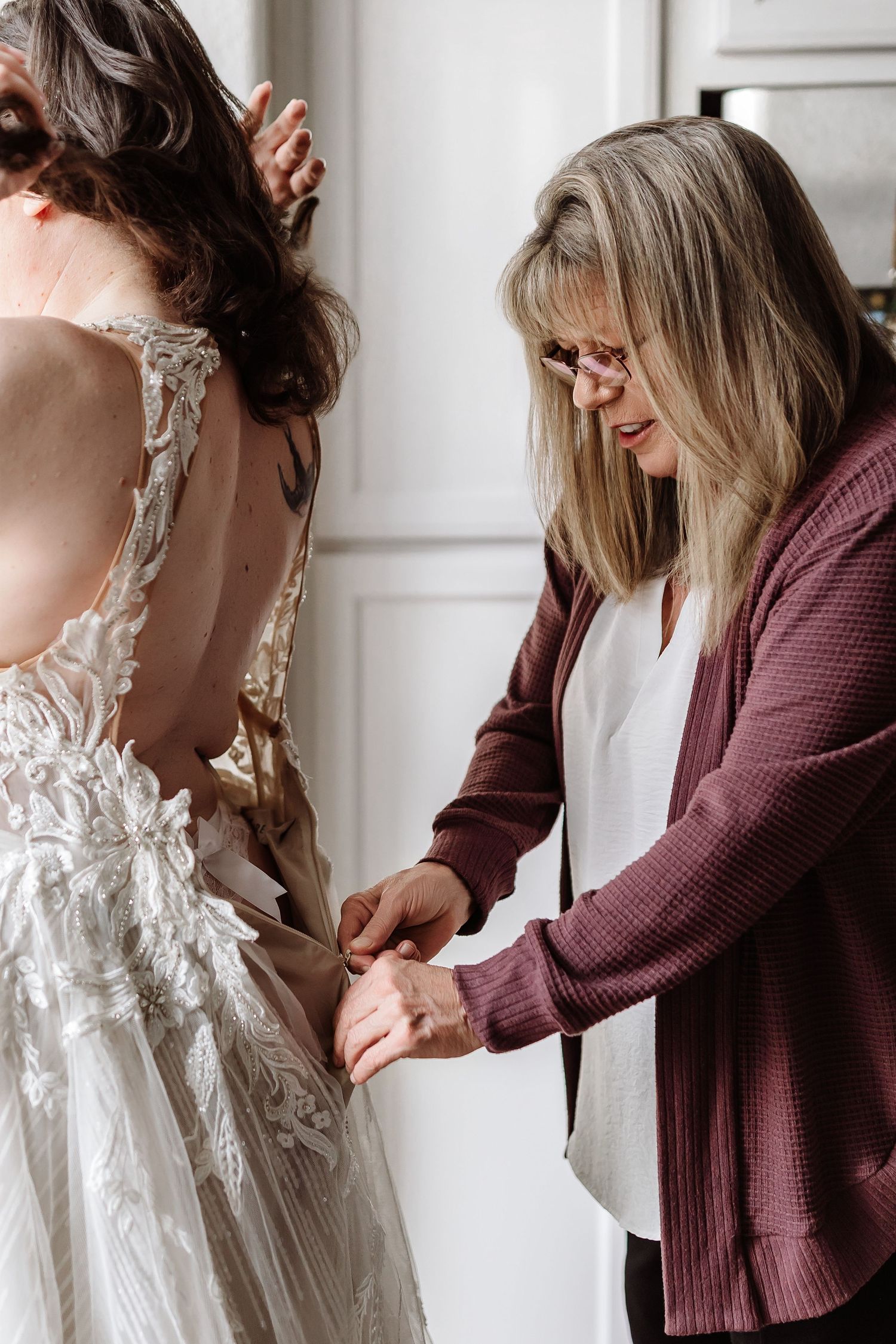 Woman in burgundy cardigan helping bride fasten her lace wedding gown from behind.