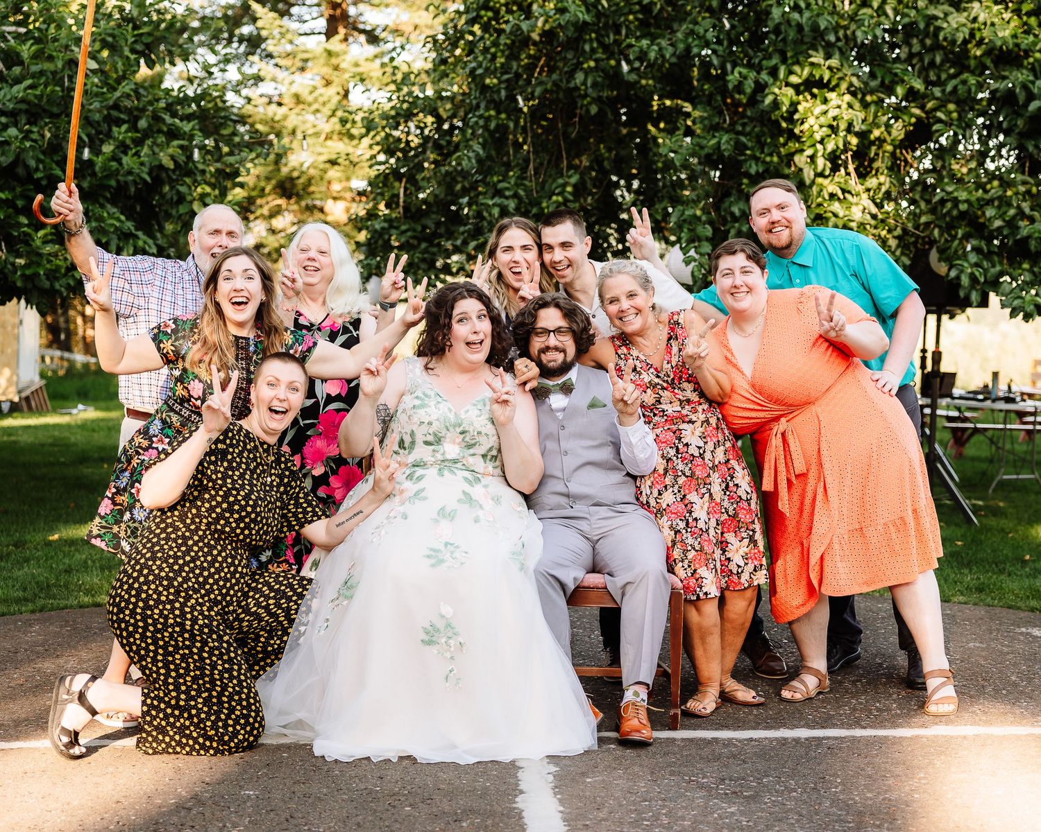 Bride and groom pose with joyful wedding party group photo outdoors in sunny garden setting.