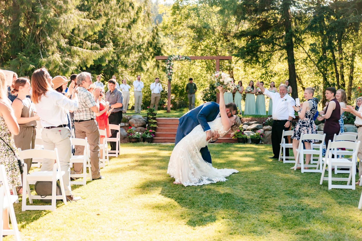 Newlyweds share a dip kiss while guests cheer along the aisle at outdoor wedding ceremony.