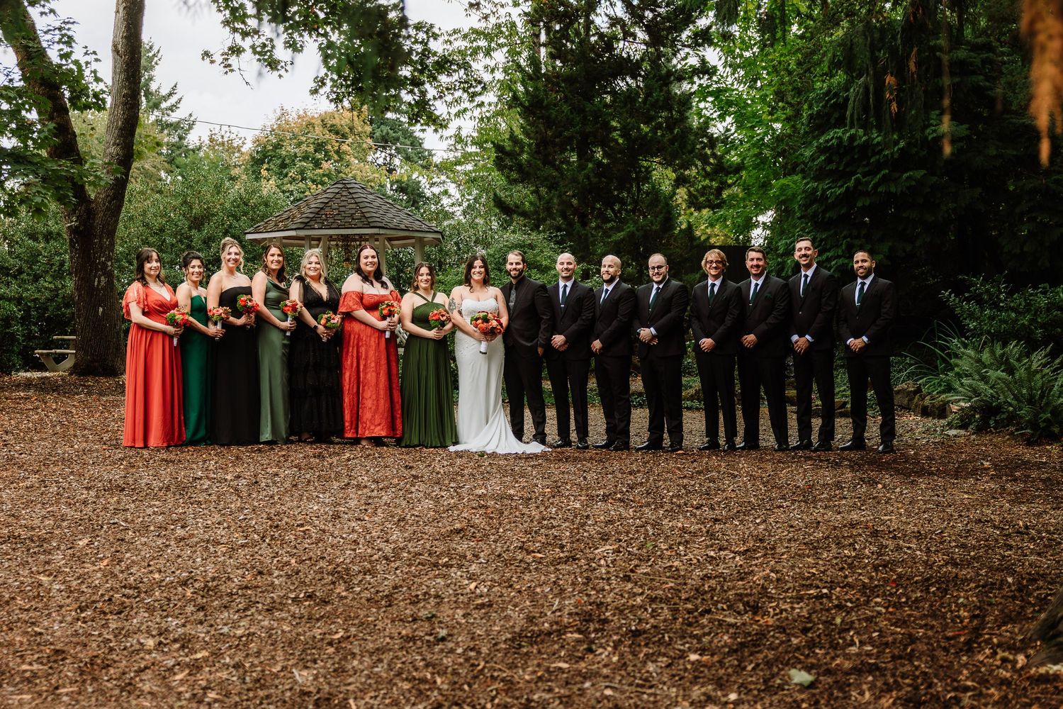 A large wedding party poses in a wooded garden with bridesmaids in red and green and groomsmen in black.