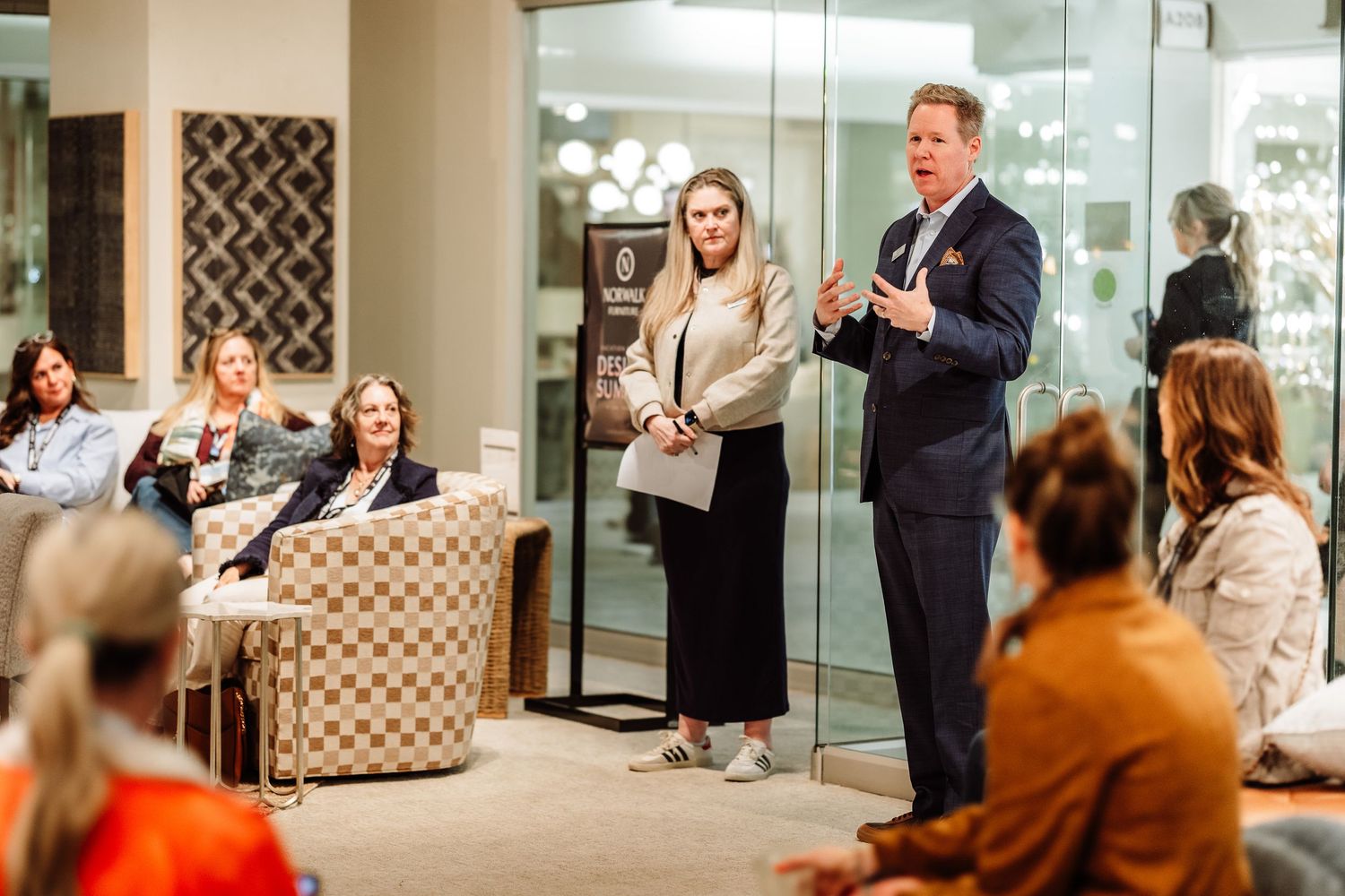 A man and woman present to a seated audience in an elegant furniture showroom.