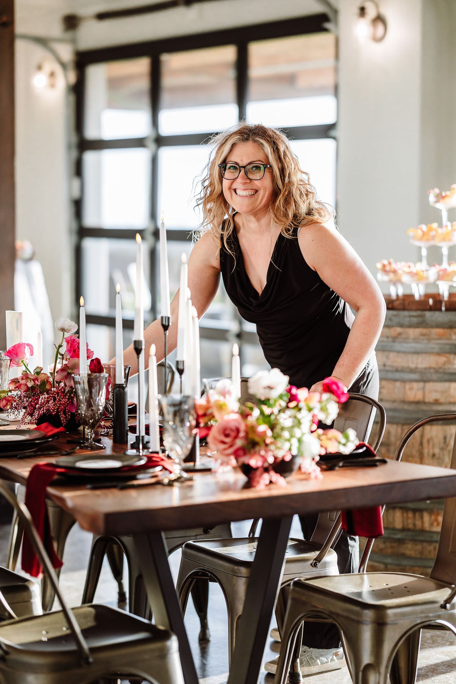 Smiling blonde woman in black dress arranging floral centerpiece at elegant event table.