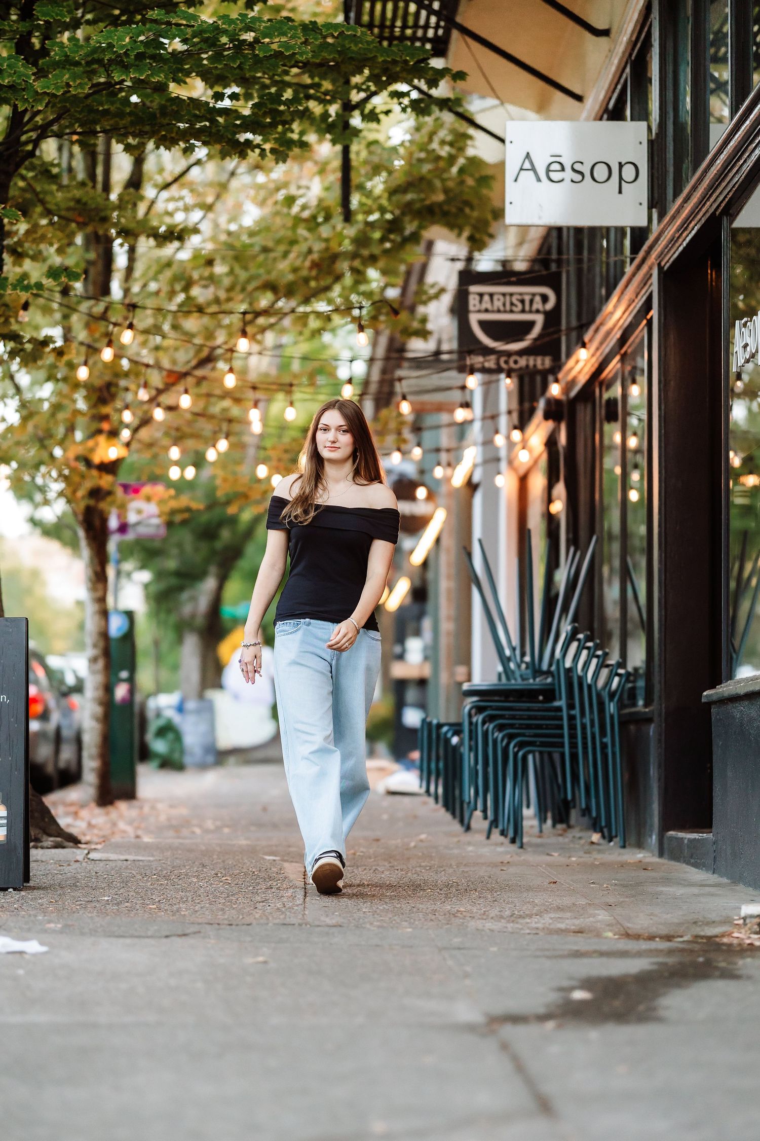 Young woman in black off-shoulder top and wide-leg jeans walking on urban sidewalk past Aesop store.