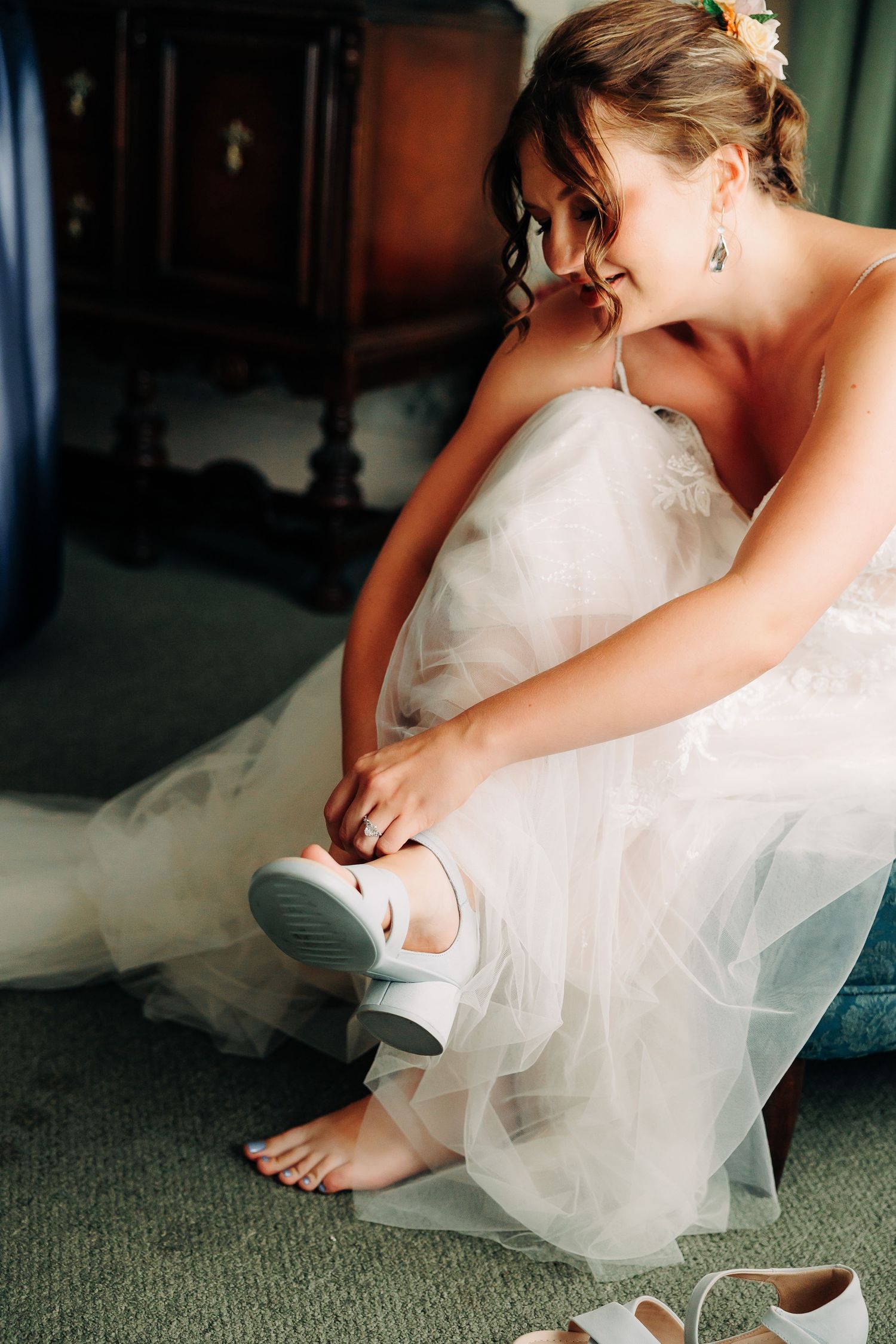 Smiling bride in white lace wedding gown putting on white bridal shoes while seated.