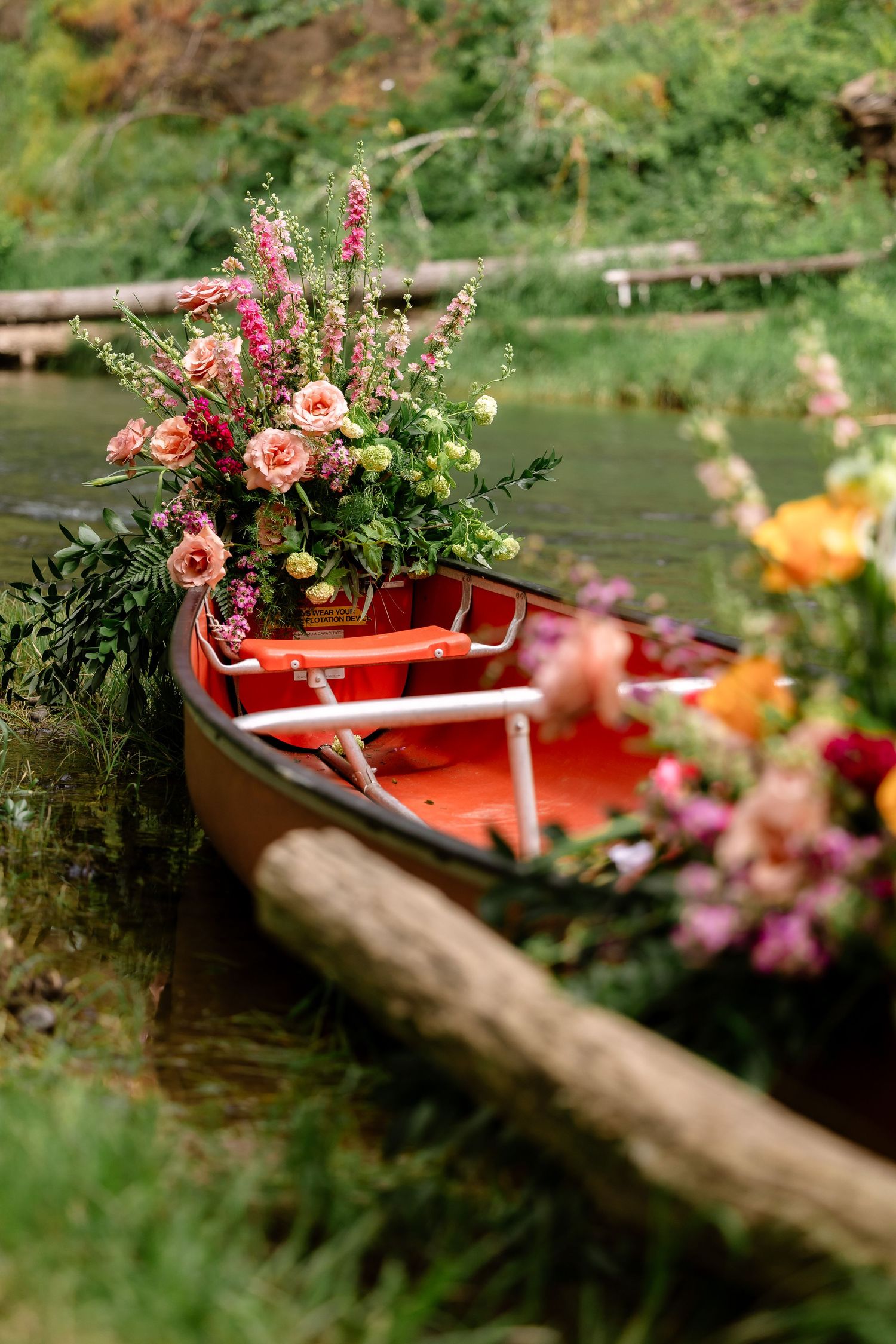Vibrant floral arrangement overflowing from a red canoe docked along a lush green riverbank at a wedding.