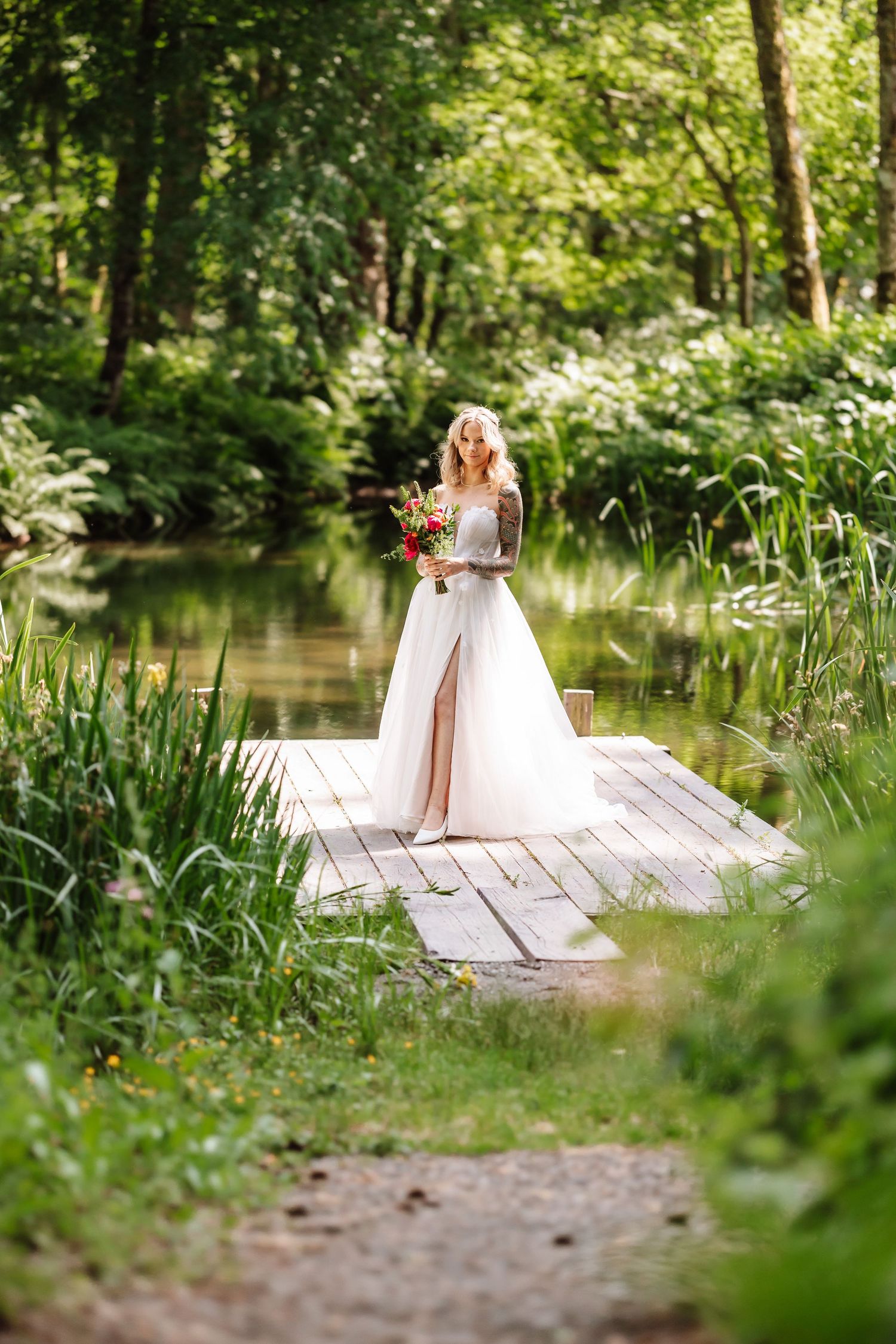 Blonde bride in lace wedding gown holds bouquet on wooden dock surrounded by lush green nature.