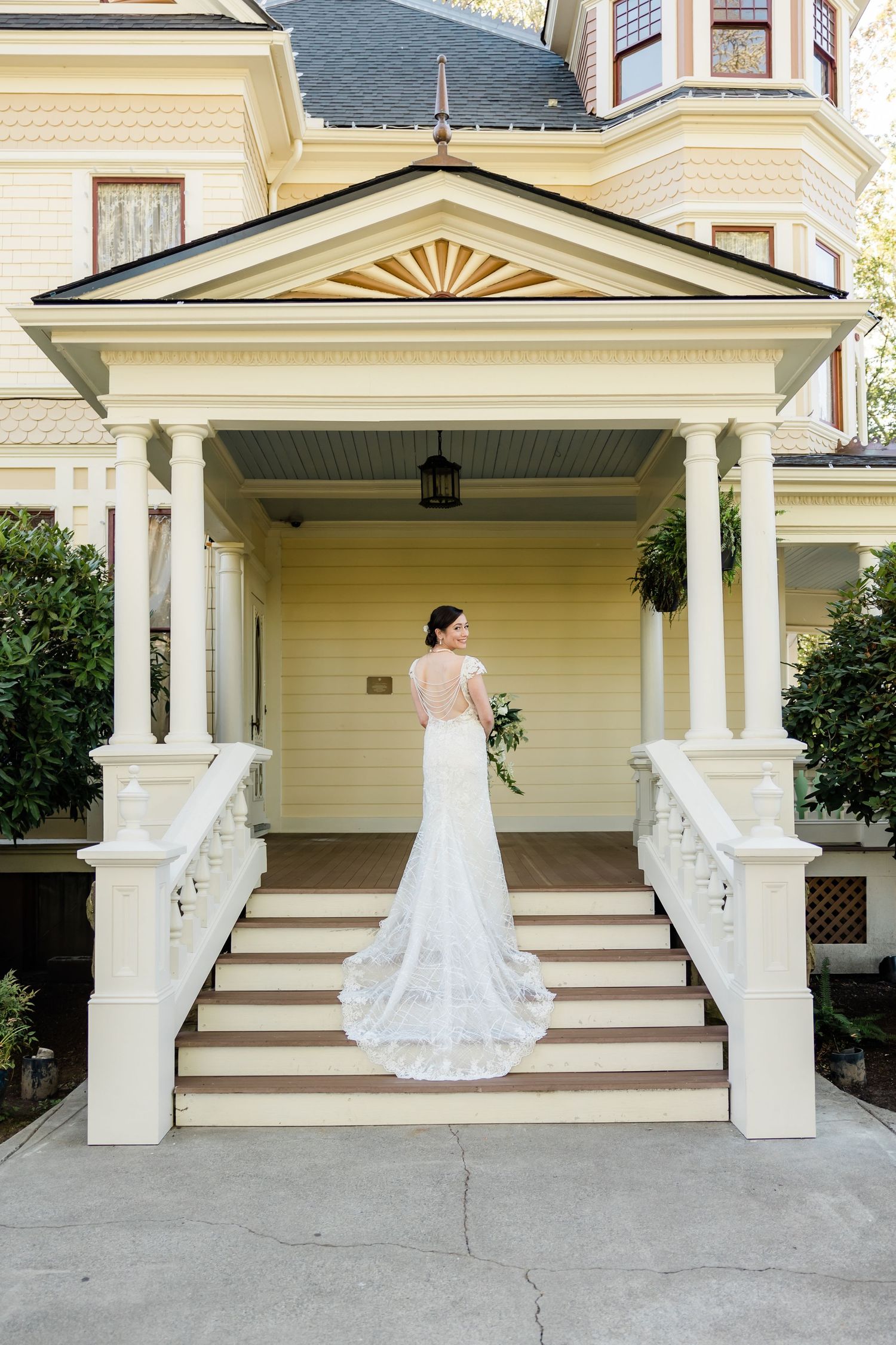 Bride in backless lace gown poses on steps of yellow Victorian mansion.