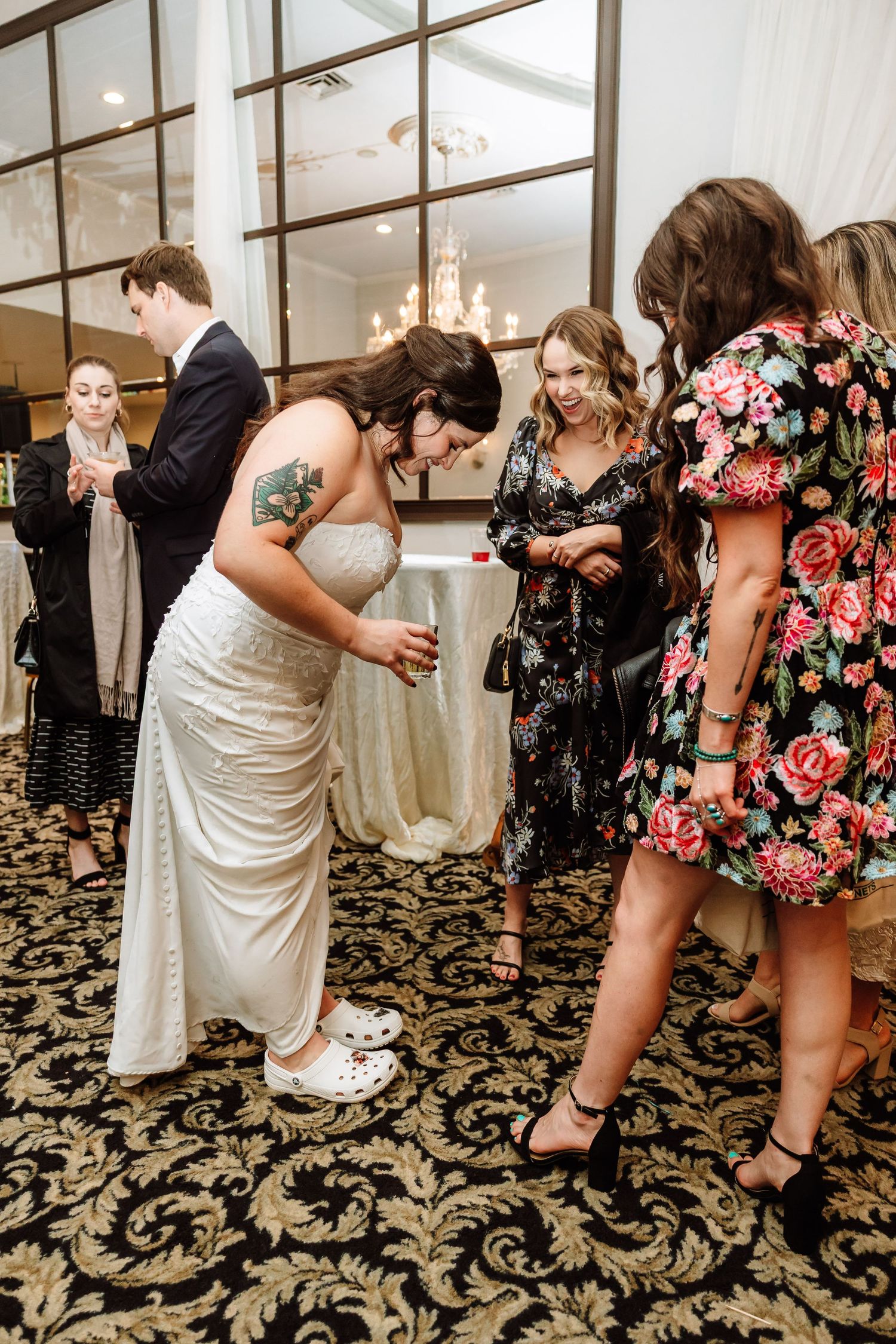 Bride with tattoos wearing white dress and Crocs showing her shoes to guests at wedding reception.