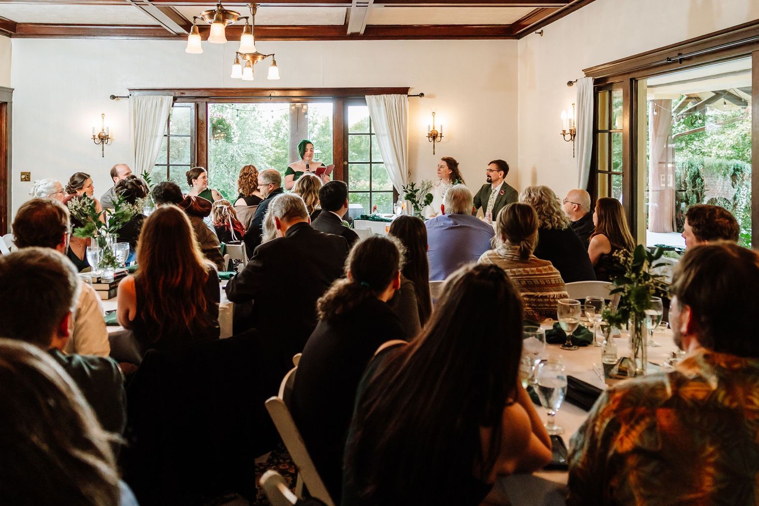 Wedding guests fill elegant reception hall as couple stands at front during speeches.