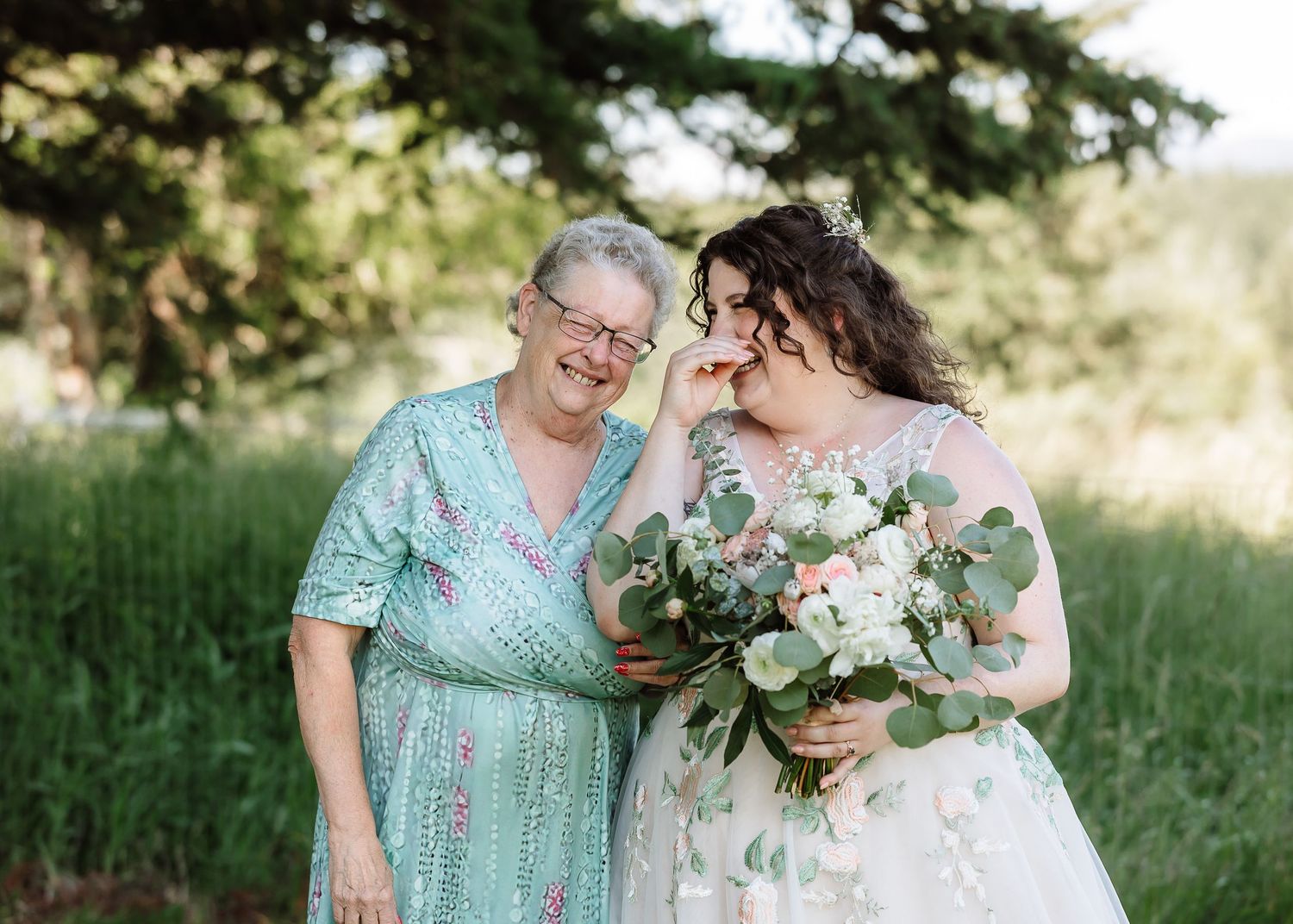 Bride in floral wedding gown laughs joyfully with older woman in green dress outdoors.